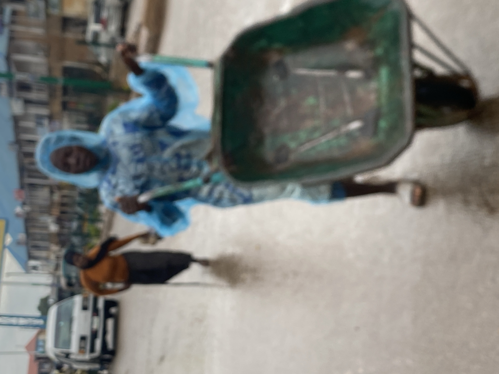 The image, which is heavily blurred and rotated counter-clockwise, captures an outdoor scene on what appears to be a paved street in an urban or semi-urban area, likely Bukuru, Nigeria.
In the foreground, a person is visible pushing a green, metal wheelbarrow. This individual is wearing a light blue or white full-body suit with a faint pattern, and their head is covered by a hood-like garment. The wheelbarrow contains some indistinct objects or tools.
Further back, another person is seen. They are dressed in a brown top and dark trousers or skirt, and they appear to be holding a long object, possibly a broom or stick. A cloud of dust near their feet suggests they are actively sweeping or engaged in a similar ground-level task.
In the background, several multi-story buildings line the street, confirming an urban setting. A white van or mini-bus is partially visible on the left side of the frame, seemingly parked or moving slowly.
The scene takes place during the daytime, characterized by bright but soft lighting, suggesting an overcast sky or perhaps early morning or late afternoon. The overall blurriness of the image prevents the identification of fine details or any discernible text. The activity depicted suggests street cleaning or general labor.