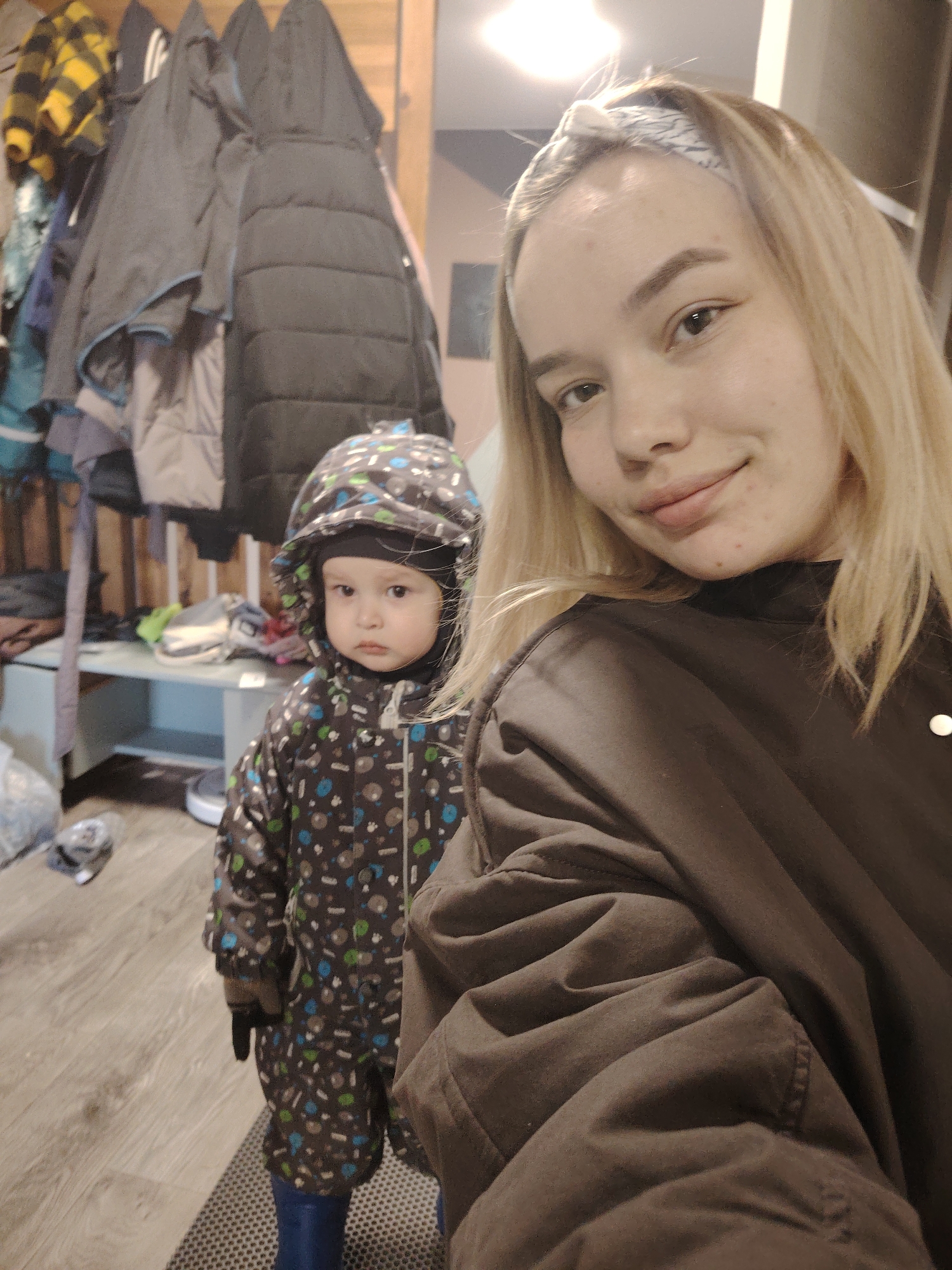 A woman with blonde hair and a patterned headband takes a selfie in what appears to be a home interior in Марковский, Russia. A young child wearing a patterned snowsuit with a hood stands to her left. The child wears gloves and blue boots. Behind the woman and child is a coat rack laden with multiple coats. A shelf with various objects on it is also visible in the background. The floor is wooden-patterned linoleum.