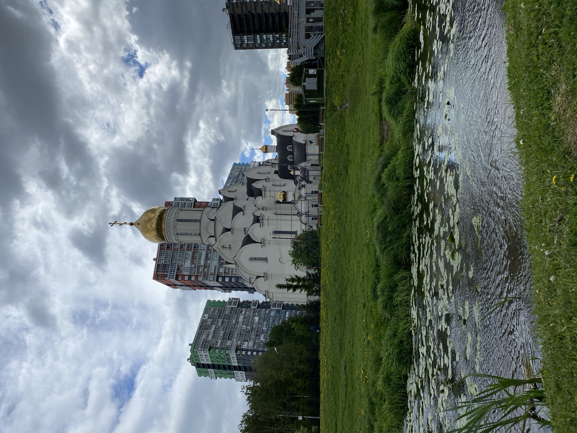 A rotated medium shot shows a white building with golden domes and crosses in front of high-rise apartment buildings, beside a grassy bank and a body of water with lily pads, under a sky of dark and light clouds in Unknown, Unknown.