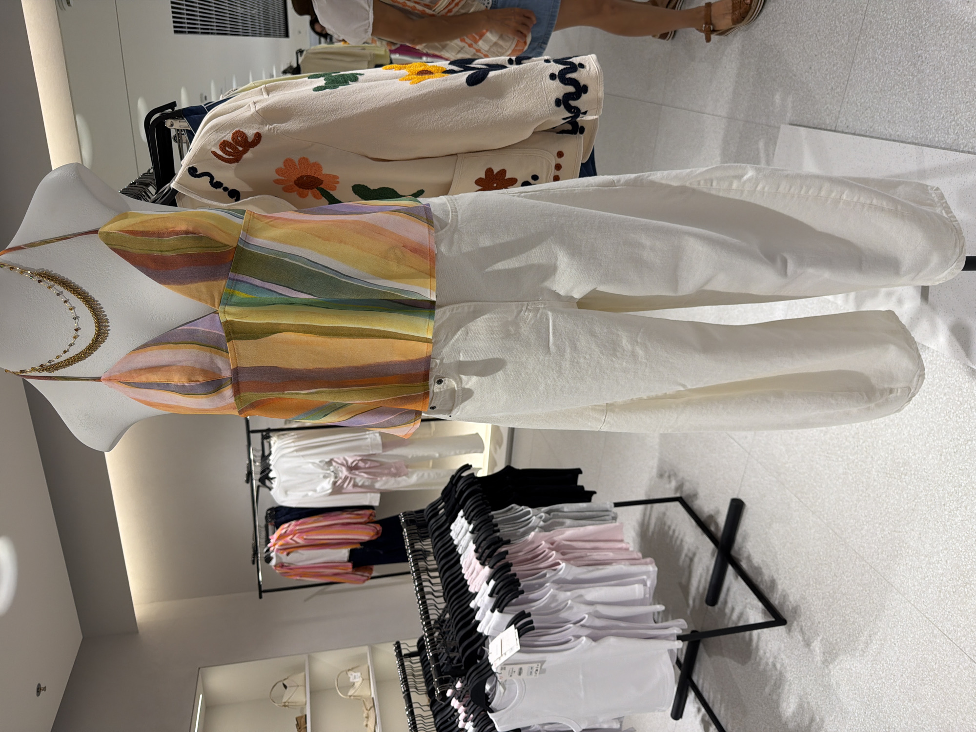 In an indoor retail environment, a headless mannequin is displayed wearing a multicolored, striped halter top and loose-fitting, cream-colored pants. The mannequin is accessorized with a layered gold necklace. To the mannequin's right, a cream-colored jacket with embroidered floral details is displayed on a hanger. In the background, racks of clothing, including white tank tops and pink blouses, are visible. White shelves displaying handbags are to the left. The floor is a light-colored speckled tile. A partial view of a person wearing a blue skirt and brown sandals is visible on the right side of the frame. The lighting appears to be artificial and diffused.