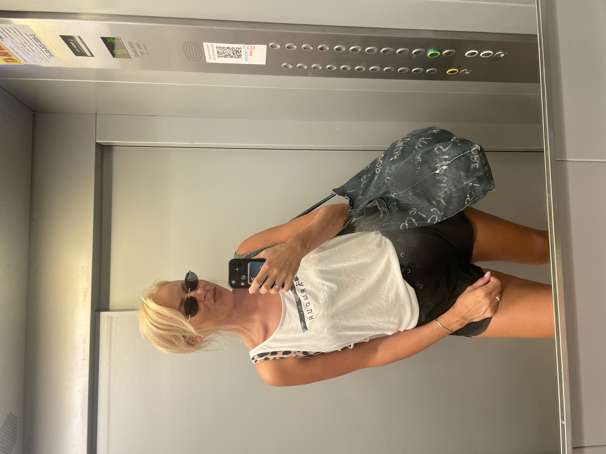 A blonde woman wearing sunglasses, a white tank top, and black shorts is taking a mirror selfie in an elevator. She is holding a smartphone in her right hand and a denim bag over her left shoulder. The elevator interior is metallic gray with a panel of buttons on the right side. The elevator is likely located in Novorossiysk, Russia, as indicated by the signage on the button panel.