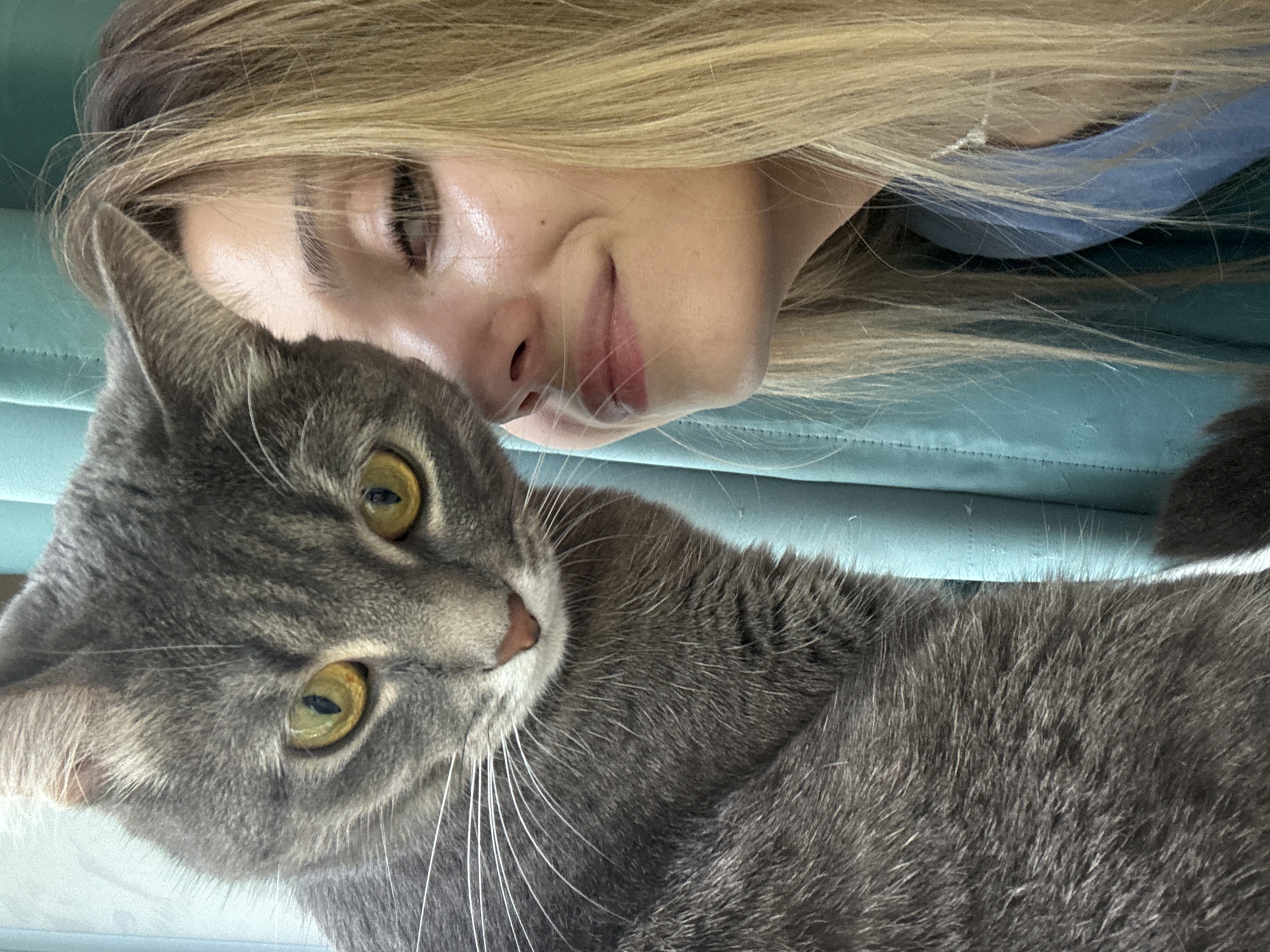 A close-up selfie shows a young woman with blonde hair and a grey tabby cat. The woman is smiling, and the cat is looking directly at the camera with yellow eyes. They are both against a teal background.