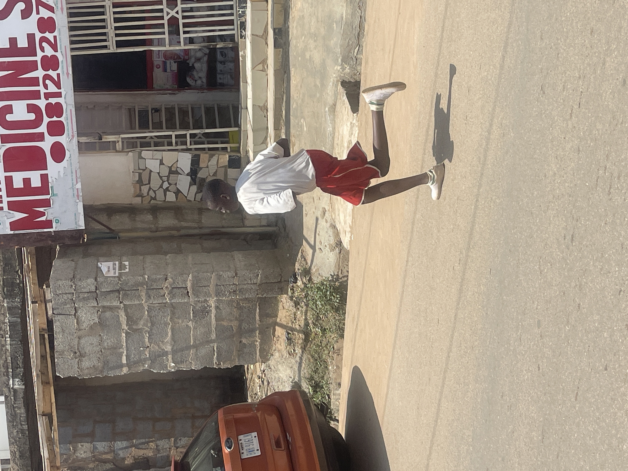 In Abuja, Nigeria, a young boy is captured mid-stride, running on a paved road. He is wearing a white t-shirt and red shorts, with white sneakers on his feet. The boy's body is angled to the right, with his right leg extended forward and his left leg bent backward, indicating he is in motion. His shadow stretches out behind him on the road.
To the left of the boy, a portion of an orange vehicle is visible, with its rear end facing the viewer. The vehicle has a license plate that reads "ABUJA BYR-916LN". Behind the boy and to the right, a building is depicted. The building has a facade made of stone blocks. Above this, a sign with red lettering is visible, reading "MEDICINE S" and a phone number "08128287". The building features a gated entrance with vertical bars. The environment appears to be an outdoor urban setting, with the road surface made of asphalt and sand. The lighting suggests it is daytime, with clear skies casting distinct shadows.
