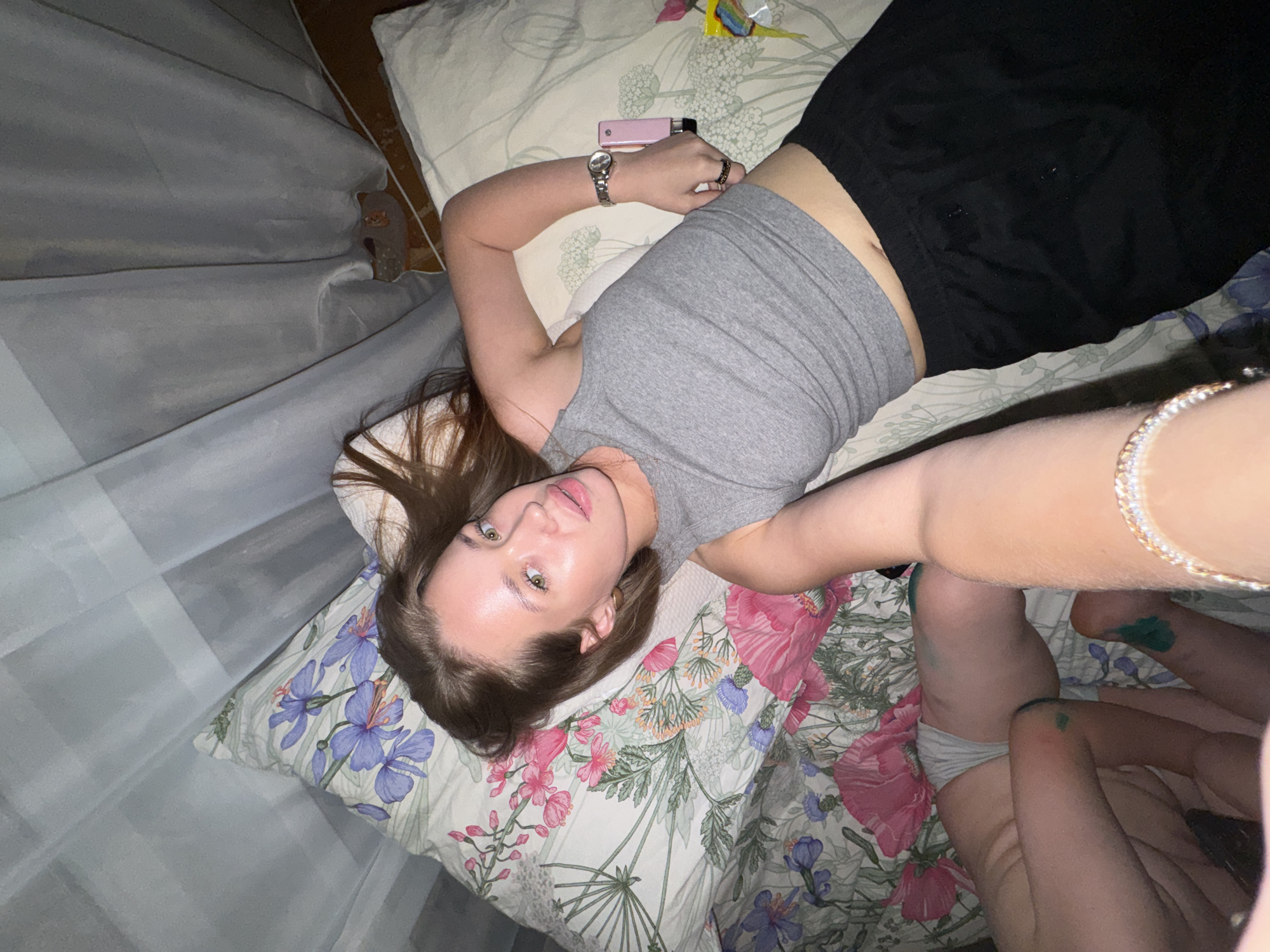 The image shows a woman lying on a bed with floral bedding. She is wearing a grey tank top and black pants. Her right arm is extended, holding a pink object, and she is wearing a watch on her left wrist. Another person's legs are visible on the left side of the image, with green paint on their knees and ankles. The background consists of sheer curtains. The location is identified as being in the city of Chaikovsky, Russia.