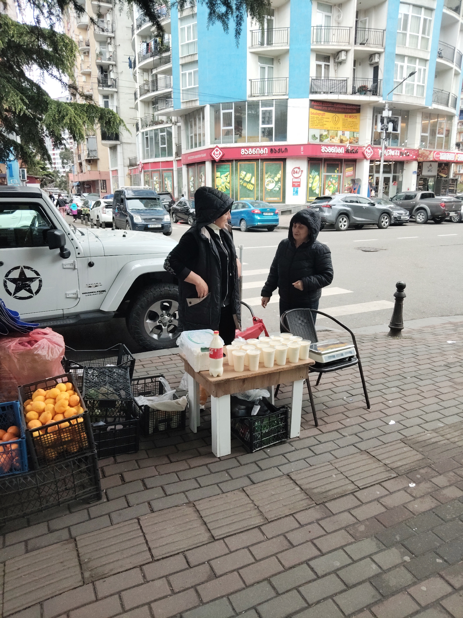 Two women stand next to a folding table covered with many cups of white liquid, a Coca-Cola bottle, and clear plastic containers in an urban Batumi, Georgia location. The women both wear dark jackets and hats and stand on a brick sidewalk next to a crosswalk. Behind them is a street with several parked cars, including a white Jeep Wrangler with a black star sticker on the door. Behind the vehicles, a multi-story building featuring both blue and beige sections stands in the background. Crates of oranges are visible on the sidewalk near the table. Signage for a "Magniti" store is seen on the corner building.