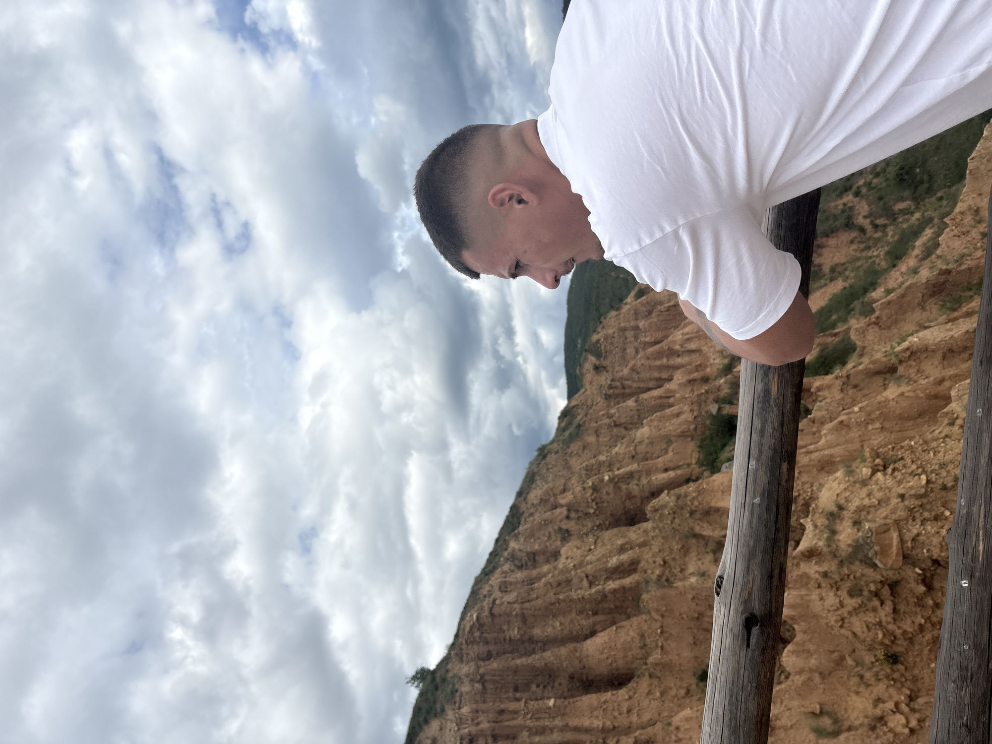 A man with a close-cropped haircut, wearing a white t-shirt, is leaning over a wooden railing overlooking a scenic view. He is looking down, towards the landscape. The background reveals a cliff face with textured rock formations and sparse vegetation. The sky is overcast with cloudy formations. The location is Stob, Bulgaria, known for its unique rock formations. The man appears to be a tourist or visitor observing the landscape.