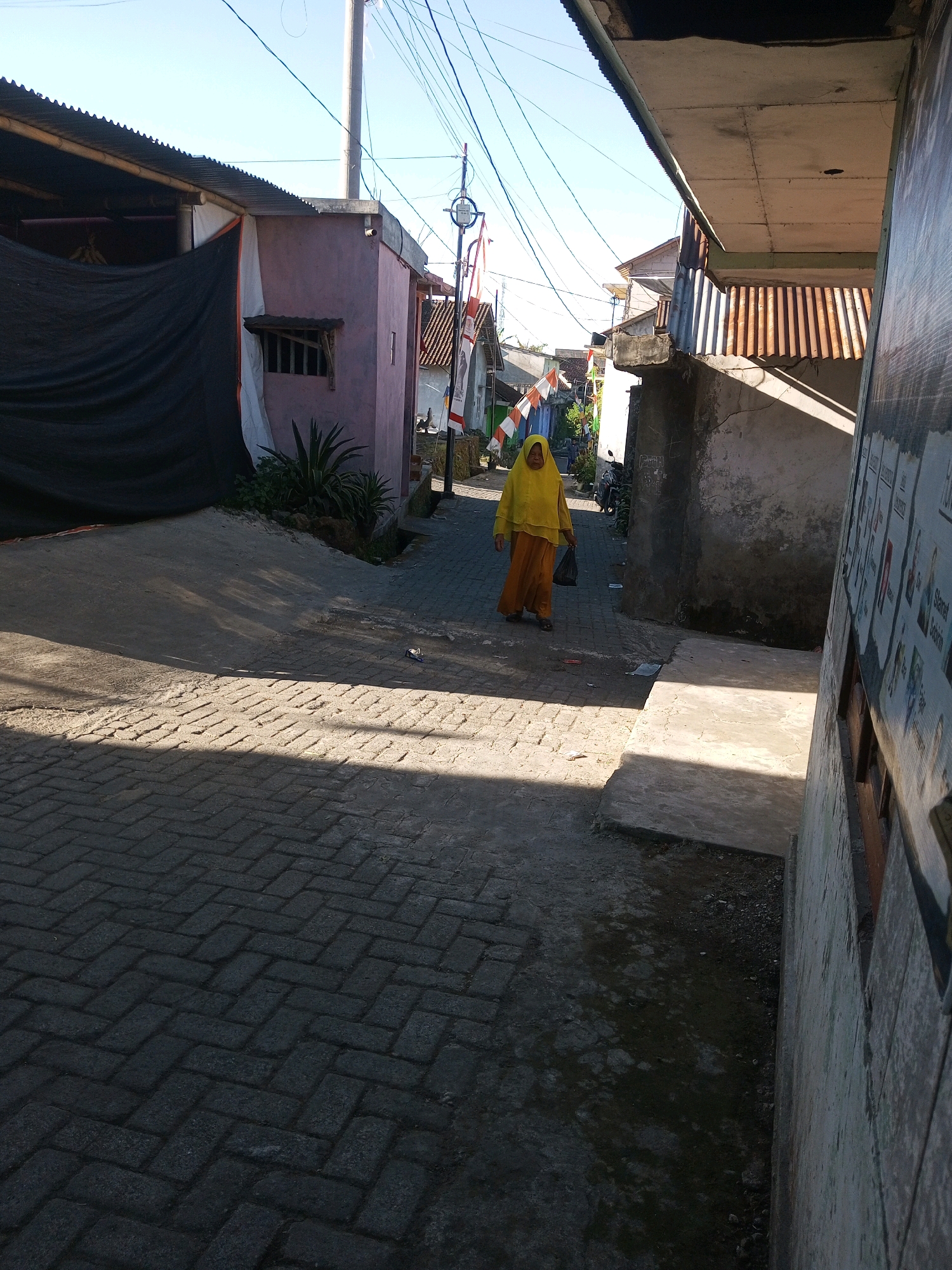 A woman wearing a bright yellow headscarf and orange skirt walks down a narrow, paved alleyway in Andongsili, Indonesia. The sun casts sharp shadows across the brick-patterned pavement. To the left, a building with a corrugated metal roof is covered in part by a large black tarp, and a small window with wooden slats is visible. Further down the alley, several Indonesian flags are displayed, indicating a festive or patriotic occasion. To the right, a wall covered in posters or flyers rises alongside the alley. The overall scene depicts a typical residential street in a village, with modest buildings and overhead electrical wires contributing to the urban environment.