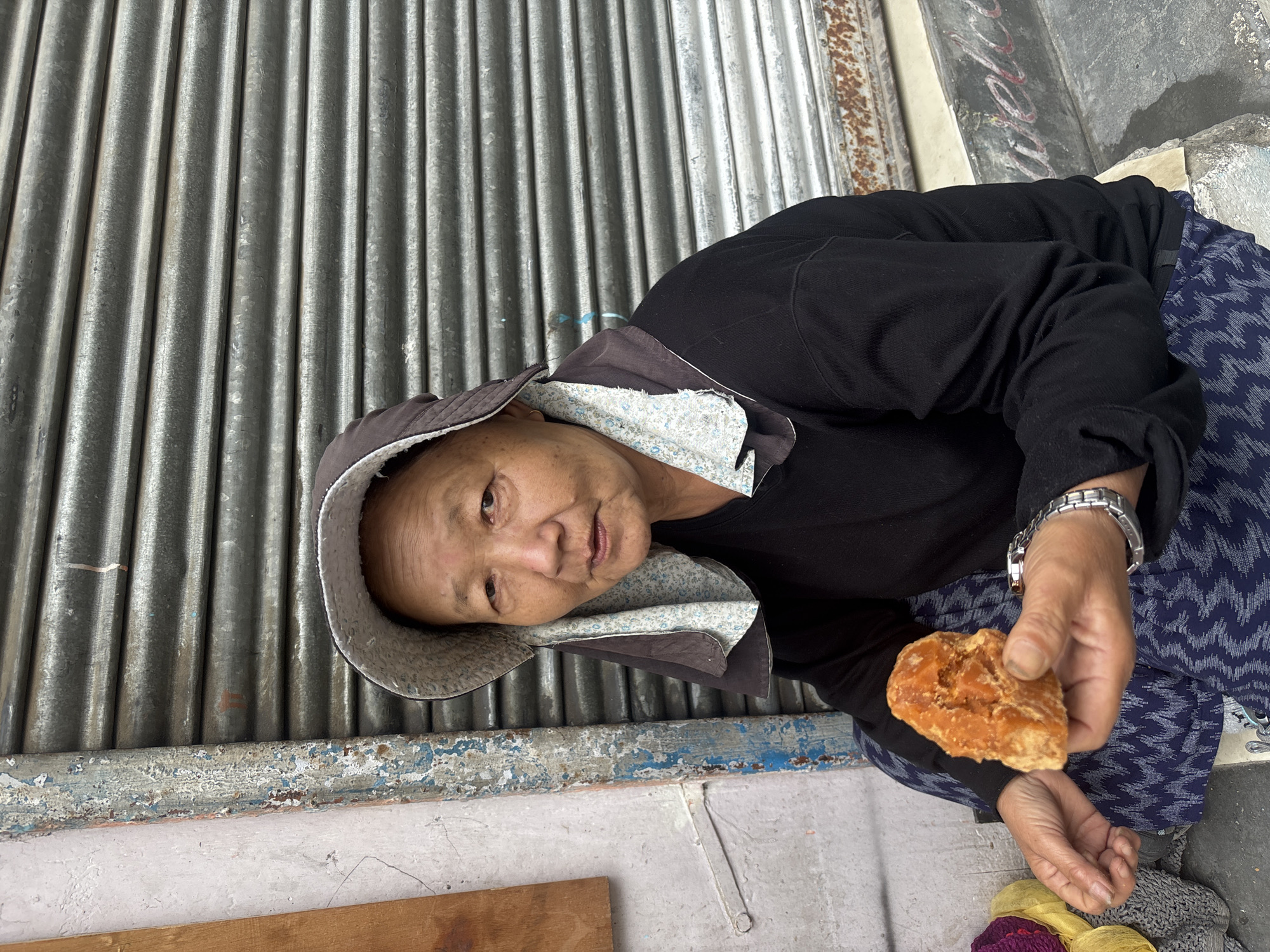 A close-up shot shows an elderly woman sitting outdoors, leaning against a corrugated metal wall. She is wearing a black hooded jacket with a floral-patterned lining and a dark blue patterned skirt. The woman holds a piece of what appears to be jaggery, a type of unrefined cane sugar, in her right hand. A silver watch is visible on her wrist. Her face is weathered, and she looks directly at the camera with a neutral expression. The background consists of the textured metal wall and a concrete surface with peeling blue paint at the bottom. The lighting suggests it could be daytime. The scene is likely in Vashisht, India, as indicated by the location context. There is no visible text in the image.