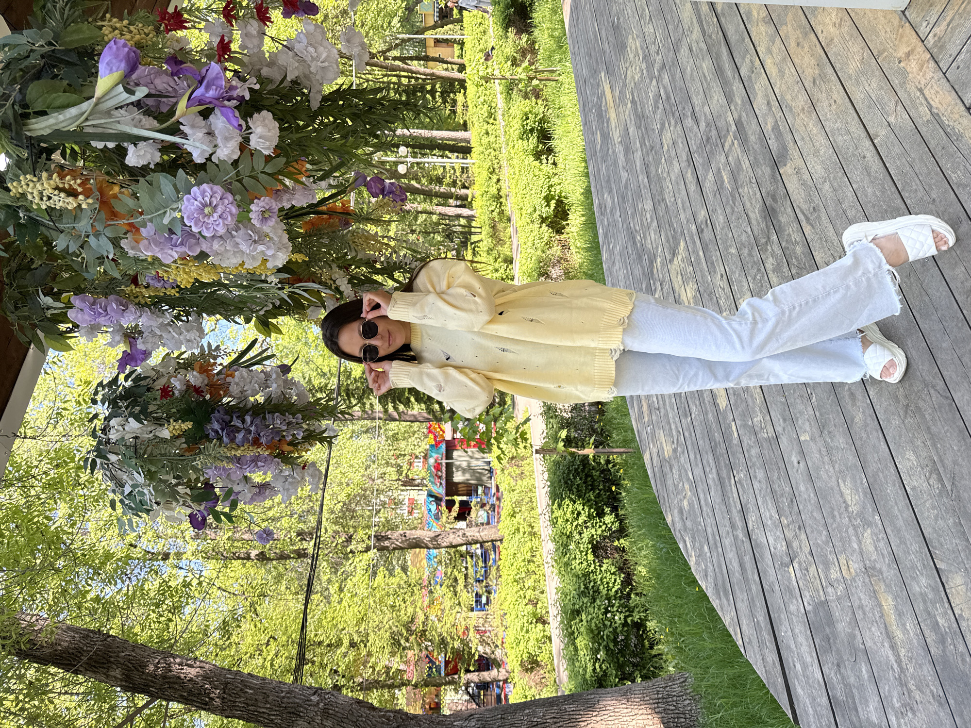 A young woman stands on a wooden deck in what appears to be a park or outdoor area in Perm, Russia. She is wearing light-colored jeans, a yellow sweater with holes, and white sandals. She has sunglasses on and is making a peace sign with both hands. A large floral arrangement hangs overhead. The background includes trees, grass, and a structure with colorful elements, possibly a play area. The scene is well-lit with natural daylight.