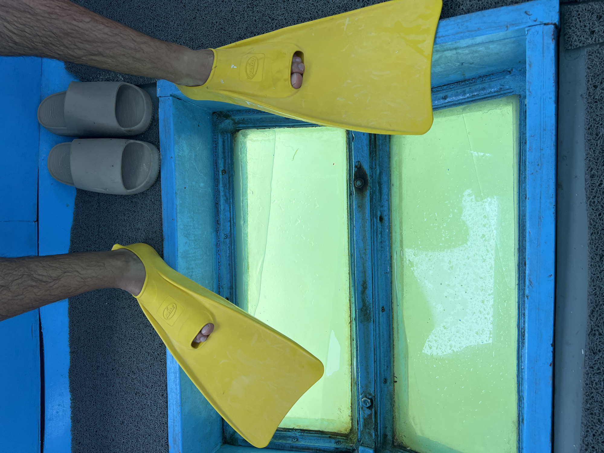 The image shows a person wearing yellow flippers standing on a glass-bottomed boat. Next to the person are a pair of gray slides. The glass in the bottom of the boat has a bluish frame and is showing water underneath. The surroundings are a textured, gray material. The location is Unknown, Indonesia.