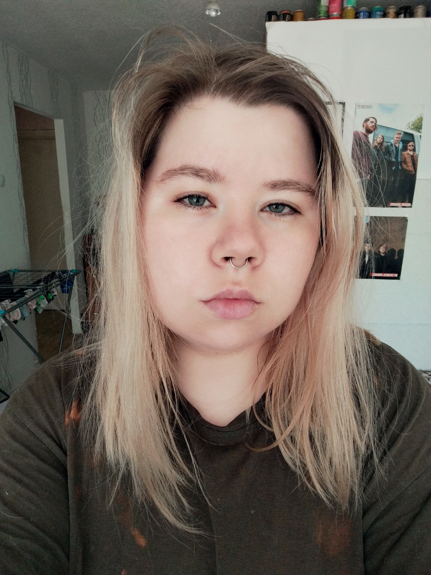 A medium shot captures a person with fair skin, light blonde hair, and blue eyes. The person has a septum piercing and is wearing an olive green t-shirt. The background includes a white wall with two posters featuring bands or groups of people. A clothes drying rack is visible in the left background and what appears to be cleaning supplies are located along the top of the wall. Location: Unknown, Unknown.