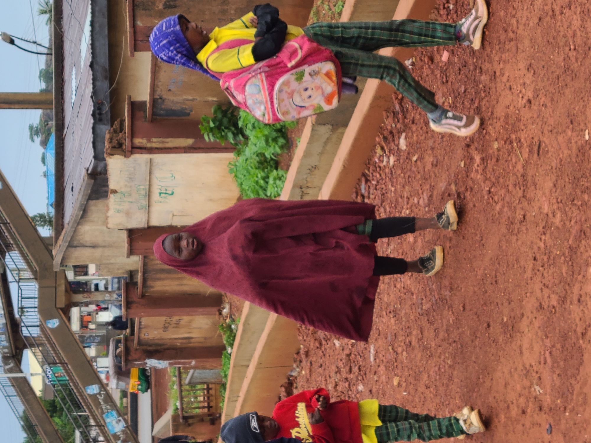 The image depicts three children walking on a dirt road in Gura Top, Nigeria. The child on the left wears a black beanie, a red sweatshirt with yellow text, and green plaid pants. The child in the center wears a maroon abaya and black leggings, and the child on the right wears a yellow shirt, black arm sleeves, a blue headscarf, and green plaid pants. The child on the right also carries a pink backpack with cartoon characters. In the background, there is a pedestrian overpass, a yellow and green auto-rickshaw, and buildings with corrugated metal roofs. The environment appears to be somewhat dilapidated, with loose soil and debris on the road. The sky is overcast.