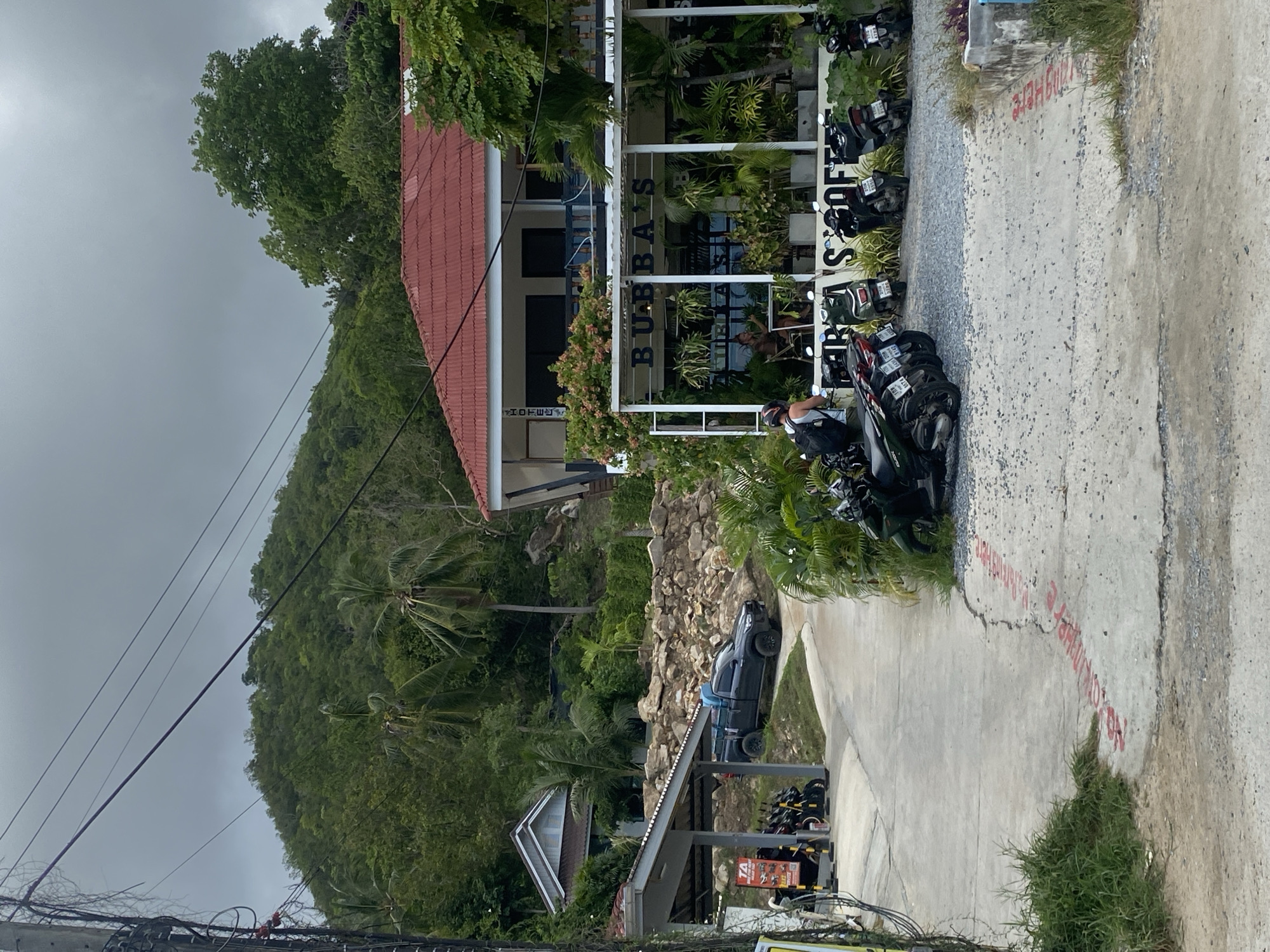 This is an outdoor scene in ตำบลเกาะพะงัน, Thailand, featuring a road leading towards a building with a red roof and an outdoor seating area for a coffee shop. Several motorcycles are parked on a gravel and concrete surface in front of the coffee shop. A black pickup truck is parked on a paved area near a covered shelter, next to a row of motorcycles. The building behind the coffee shop has "HOTEL" written vertically on its side. A person with a backpack is standing near the motorcycles, facing the coffee shop. The background consists of a steep, densely vegetated hillside with palm trees. Overhead, electrical wires are visible against a cloudy sky. The ground is a mix of concrete, gravel, and some grassy areas, with red painted markings on the pavement.