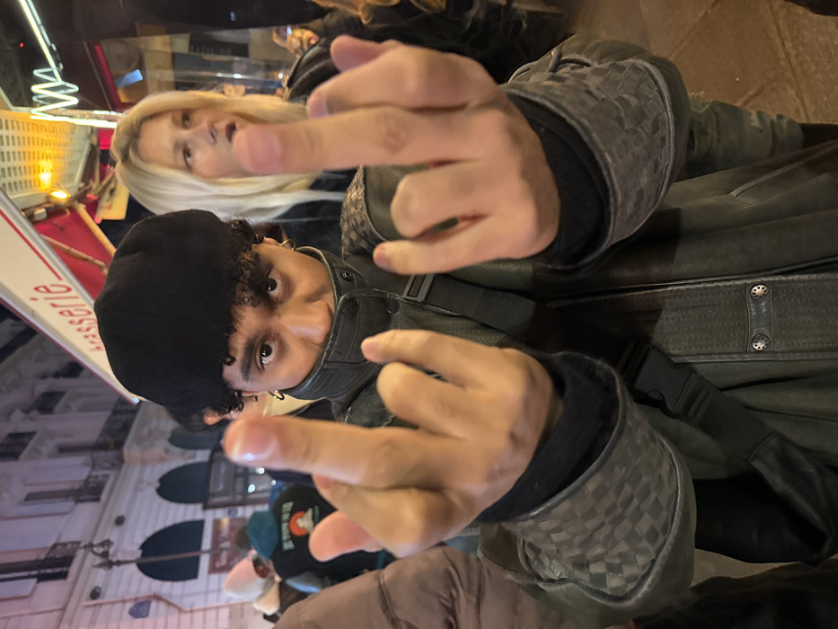 A person with curly hair, wearing a black beret and a dark jacket with a checkered pattern on the sleeves, is giving the middle finger to the camera with both hands. A woman with blonde hair is standing behind them. The background includes a building with rounded windows, and a red and white sign with "Brasserie" written on it. The overall scene appears to be taken outdoors at night.