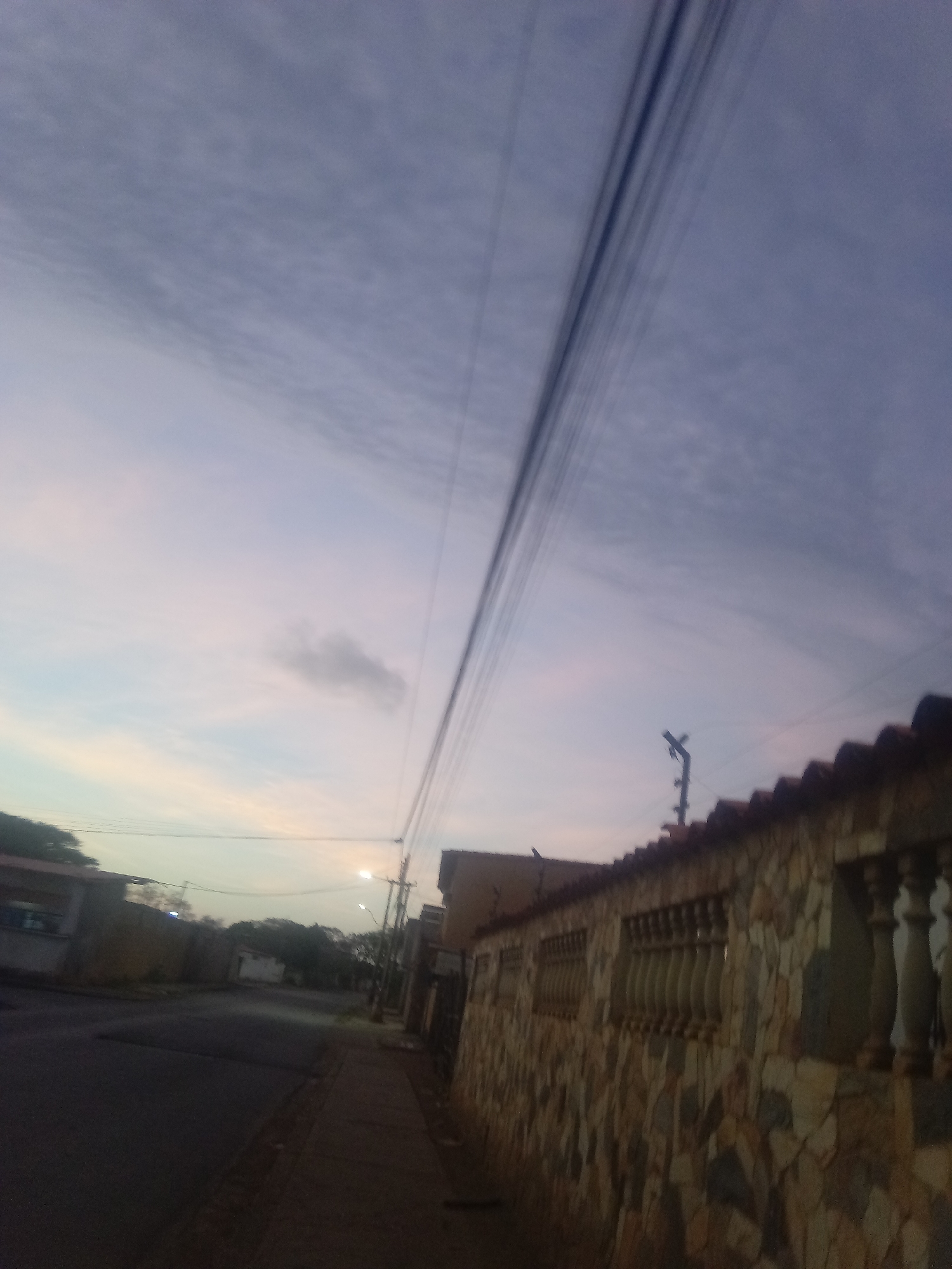 The image depicts a street scene in Juan Griego, Venezuela, at dusk or dawn. The sky is a mix of light blues, purples, and pinks, with streaks of clouds. Power lines stretch across the top of the frame, angling downward from left to right. On the right side of the image is a stone wall with a red tile roof and decorative balusters. The street and sidewalk are visible on the left, with buildings and trees in the background. A streetlight illuminates the scene in the distance. There are no people visible in the image.