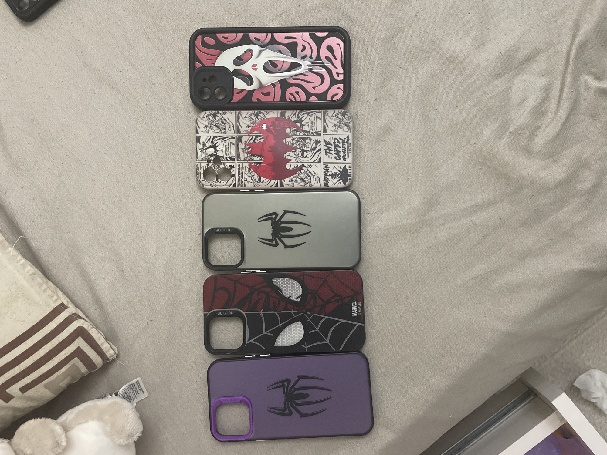 The scene, situated in the city of Ihtiman, Bulgaria, features five smartphone cases arranged in a horizontal line on a light beige, textured fabric surface. From left to right, the cases are: a purple model with a black spider emblem; a black and red case depicting a split Spider-Man mask design, marked "MARVEL" and "SO COOL"; a grey case with a black spider emblem, also marked "SO COOL"; a white case illustrated with black and white Batman comic panels and a central red Batman logo, bearing text such as "BATMAN" and "THE CAPED CRUSADER!"; and a black case displaying a pink and black melting smiley face pattern beneath a white, dripping Ghostface mask. Each case incorporates a protective bumper, either black or purple.
In the upper left quadrant, a segment of a cream-colored, fluffy stuffed animal with a visible white tag is present, positioned beside a beige pillow adorned with a brown geometric pattern. The underlying fabric surface, consistent with upholstery or bedding, shows subtle creases and a fibrous texture. A white rectangular object with a silver edge, possibly a frame or book, is partially visible in the lower left corner, near a small piece of white material. A small portion of a dark object is also visible in the upper right corner.
