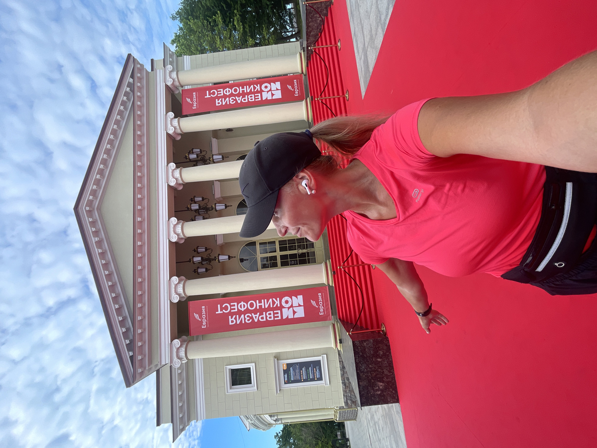 The image shows a woman wearing a bright pink t-shirt, a black baseball cap, and a black fanny pack. She is standing on a red carpet in front of a building with large white columns and banners. The banners are red and have white text that appears to say "Евразия Кинофест". This event is likely taking place at the "Sochi State Circus" in Sochi, Russia, a venue known for hosting cultural events. The woman is looking up and to her right, possibly posing for a photo or looking at something off-camera. The scene is outdoors, under a cloudy sky, with trees visible in the background. The red carpet suggests a formal event, possibly a film festival or premiere.