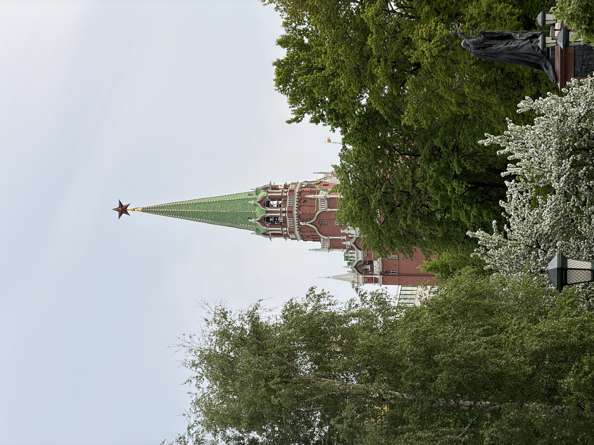 The image shows the Moscow Kremlin, specifically one of the towers topped with a red star. The tower is framed by trees with green foliage, and white flowering trees are visible to the right. The sky is overcast. A statue is partially visible at the upper right corner of the frame.