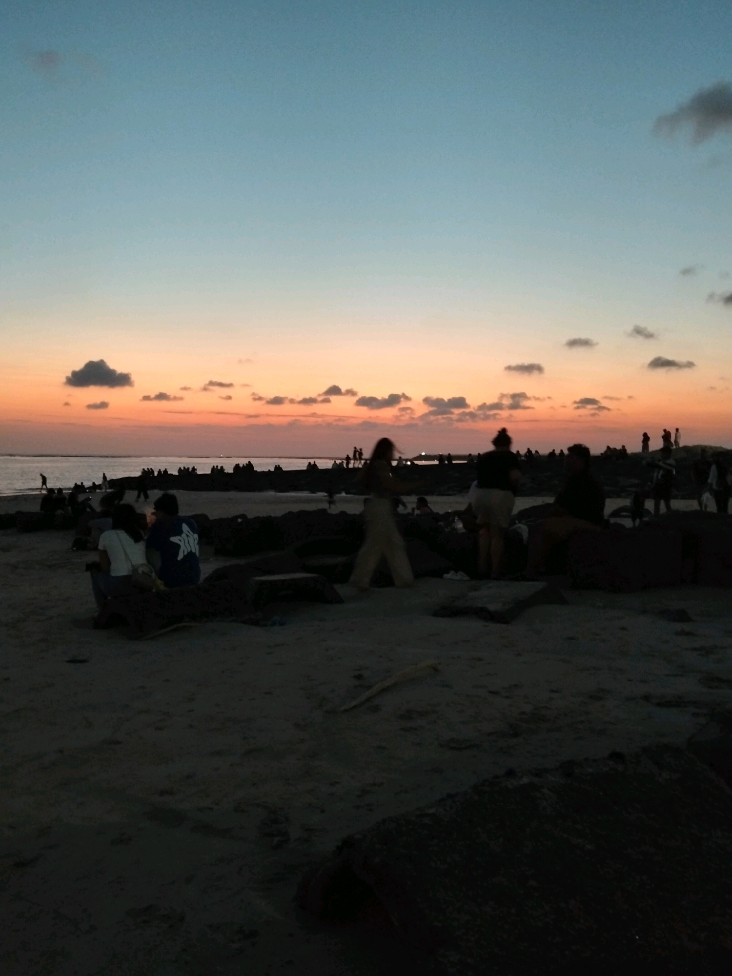 This is a silhouetted, medium-shot photograph taken at dusk on the beach in Kuta, Bali, Indonesia. The sky transitions from a pale blue at the top to a gradient of orange and pink hues near the horizon, with scattered, dark clouds. In the foreground, dark sand is visible, with several large rocks. A few individuals and small groups of people are scattered across the beach and along a rocky jetty that extends into the sea. The people are depicted in silhouette, with their forms indistinct. Some are sitting on the sand, while others are standing or walking. The sea is visible in the background, reflecting the colors of the sunset. The overall atmosphere is serene and peaceful, characteristic of a sunset gathering on a popular beach.