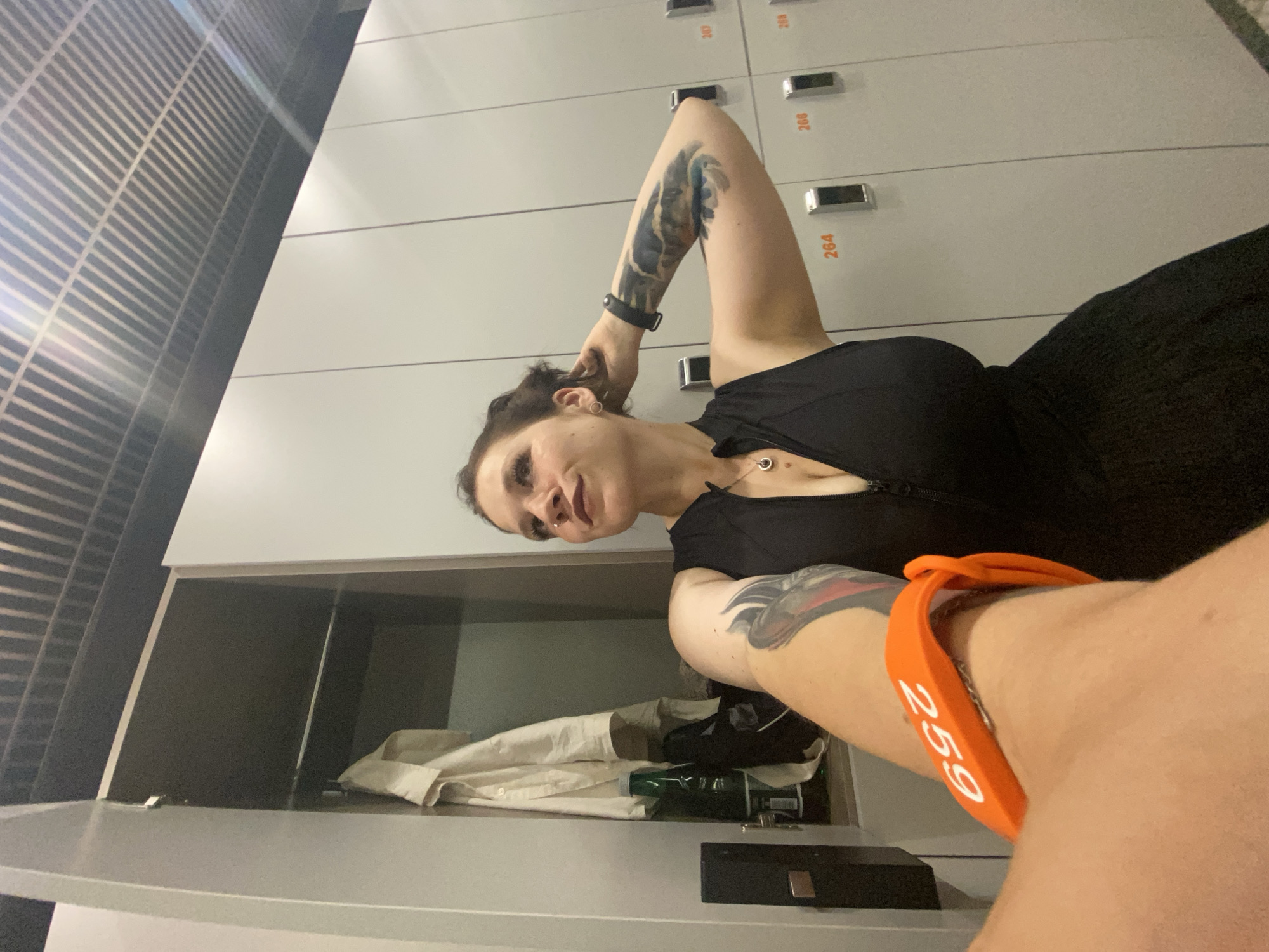 A woman with dark hair and tattoos on her arms poses in a locker room. She is wearing a black top and has an orange wristband with the number "259" on it. The background shows a row of gray lockers with numbers such as "264," "266," and "267." An open locker in the foreground contains some white fabric and a green bottle. The lighting suggests it might be indoors, possibly at a gym or spa, and the overall atmosphere is casual and relaxed.
