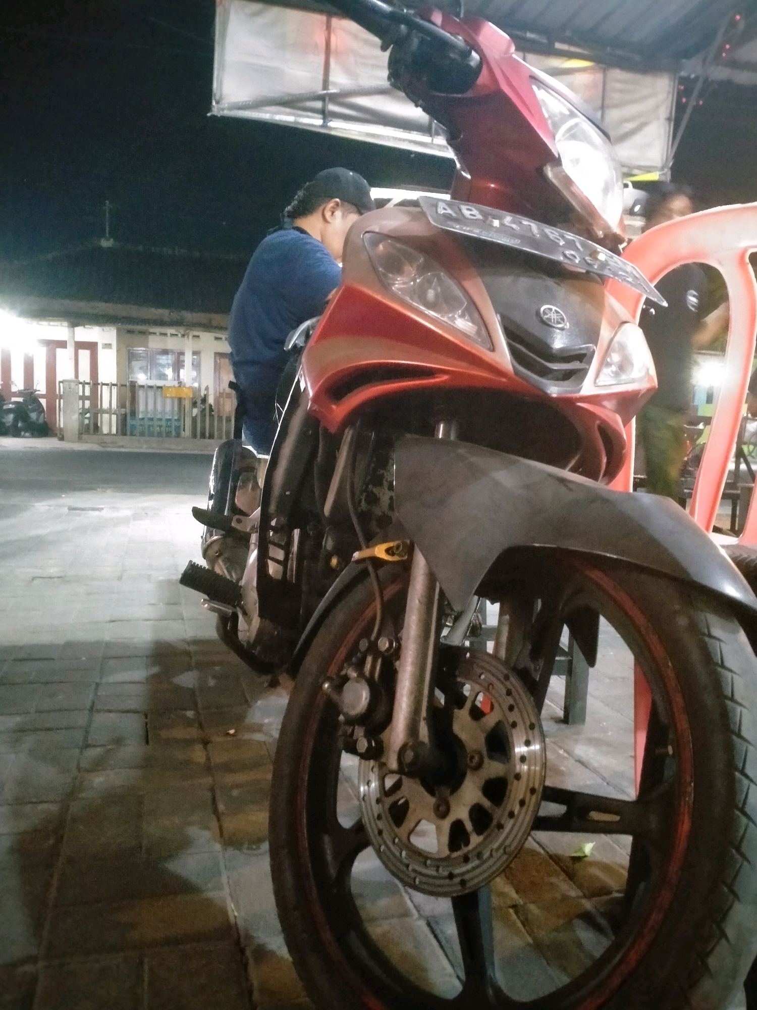 A low-angle, medium shot captures a red and black motorcycle parked on a paved walkway at night in Kasihan, Indonesia. A person wearing a blue t-shirt and a black cap is seated on the motorcycle, facing away from the camera and towards the left. The motorcycle's license plate, bearing the inscription "AB 4767 KG", is visible on the front. The background shows a dimly lit building with lights and a fence, suggesting a commercial or residential area. To the right, a portion of a red plastic chair is visible. The overall lighting is dim, with the primary light source appearing to be from the front of the motorcycle and ambient lighting from the surroundings. The walkway appears slightly wet, possibly from recent rain.