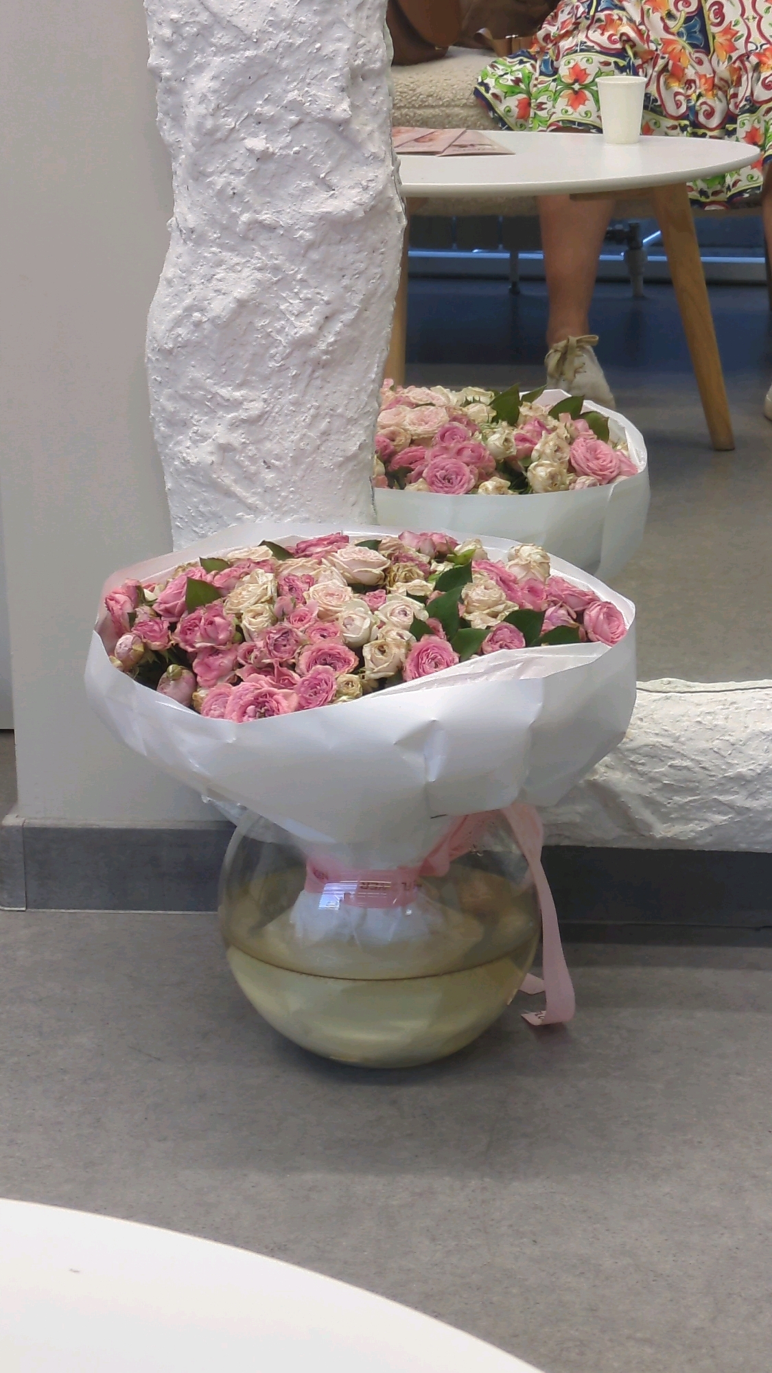 A low-angle, medium shot captures two large bouquets of pink and white roses. The bouquet in the foreground is in a clear glass vase, tied with a pink ribbon, and is positioned on a gray concrete floor. In the background, another bouquet of roses is visible, resting on the floor behind a textured white pillar. A white table with people sitting at it is visible in the far background, with a white cup and patterned clothing. The overall scene suggests an indoor setting, possibly a venue or a home, in Chișinău, Moldova.
