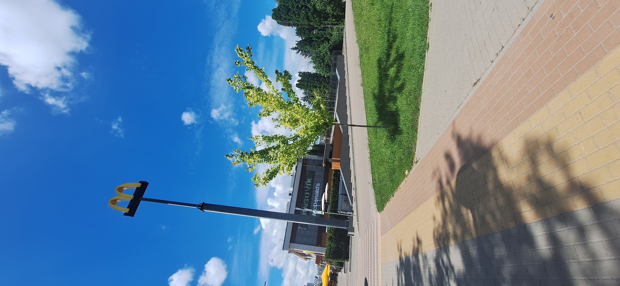 The image shows an outdoor scene with a McDonald's restaurant visible in the background. A tall pole with the golden arches logo is to the left of the McDonald's building. The building itself is modern, with large windows and a sign that reads "MCDONALD'S". A small, leafy tree stands in front of the building, casting a shadow on the grassy area. A paved walkway with a brick pattern runs diagonally across the foreground, with shadows from trees and possibly the McDonald's sign visible on it. The sky is blue with scattered white clouds. The scene appears to be a bright, sunny day. The location is identified as Bila Tserkva, Ukraine.