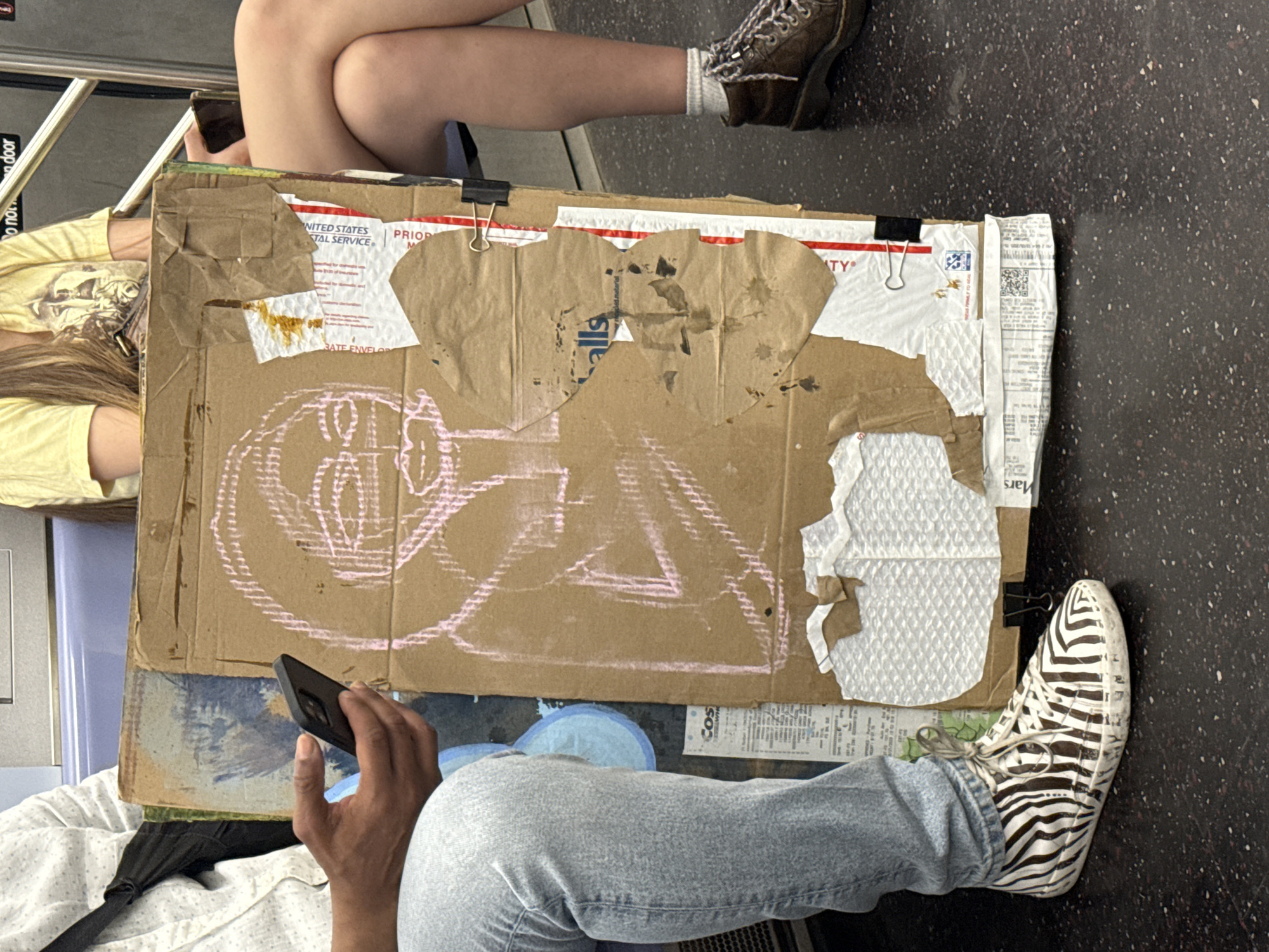 The image shows a scene on a subway car in New York City. Two individuals are seated, their legs visible. One person wears brown boots and white socks, the other wears zebra-striped sneakers and blue jeans. A piece of cardboard rests across their laps, decorated with pink chalk drawings and various paper scraps. One person is holding a black smartphone. The floor of the subway car is visible.