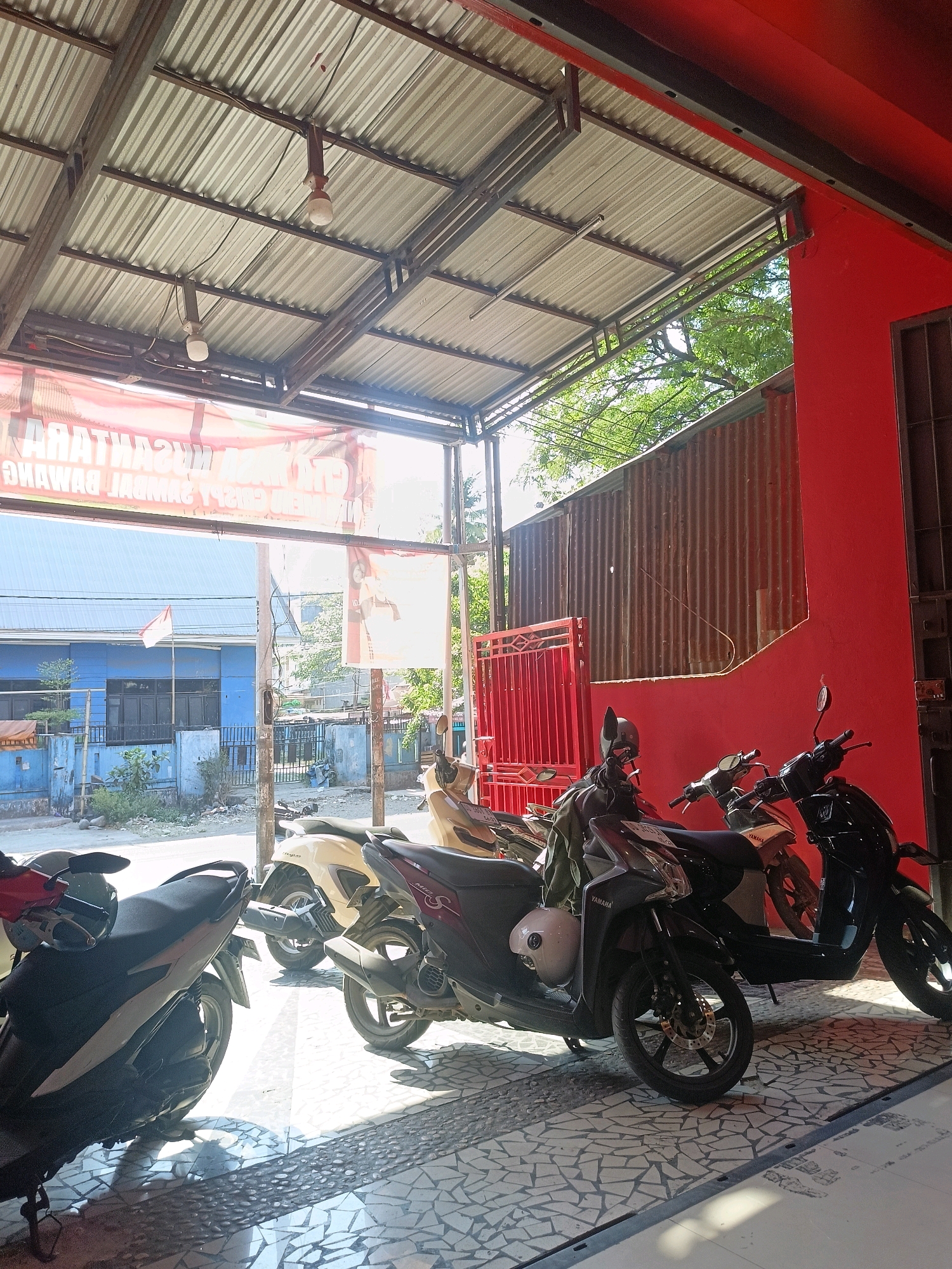 In Makassar, Indonesia, under a corrugated metal roof supported by metal beams, several motorcycles are parked in an open-air establishment. The scene is illuminated by natural light filtering through the roof and from an unseen source to the left, casting shadows on the tiled floor. Two bare light bulbs hang from the ceiling.
To the left, a light blue building with dark windows is visible, featuring a small Indonesian flag flying from a pole. A red gate stands to the right, against a weathered corrugated metal wall. A bright red wall of the establishment is prominent on the right side of the frame.
The motorcycles include a dark colored scooter in the foreground on the left, a cream colored scooter behind it, and a black scooter further to the right, with another dark colored scooter to its right. Some motorcycles have helmets and bags on them. The overall impression is a typical roadside establishment in a tropical climate, likely a shop or repair service for motorcycles. The atmosphere is casual and functional, with evidence of everyday commerce.
