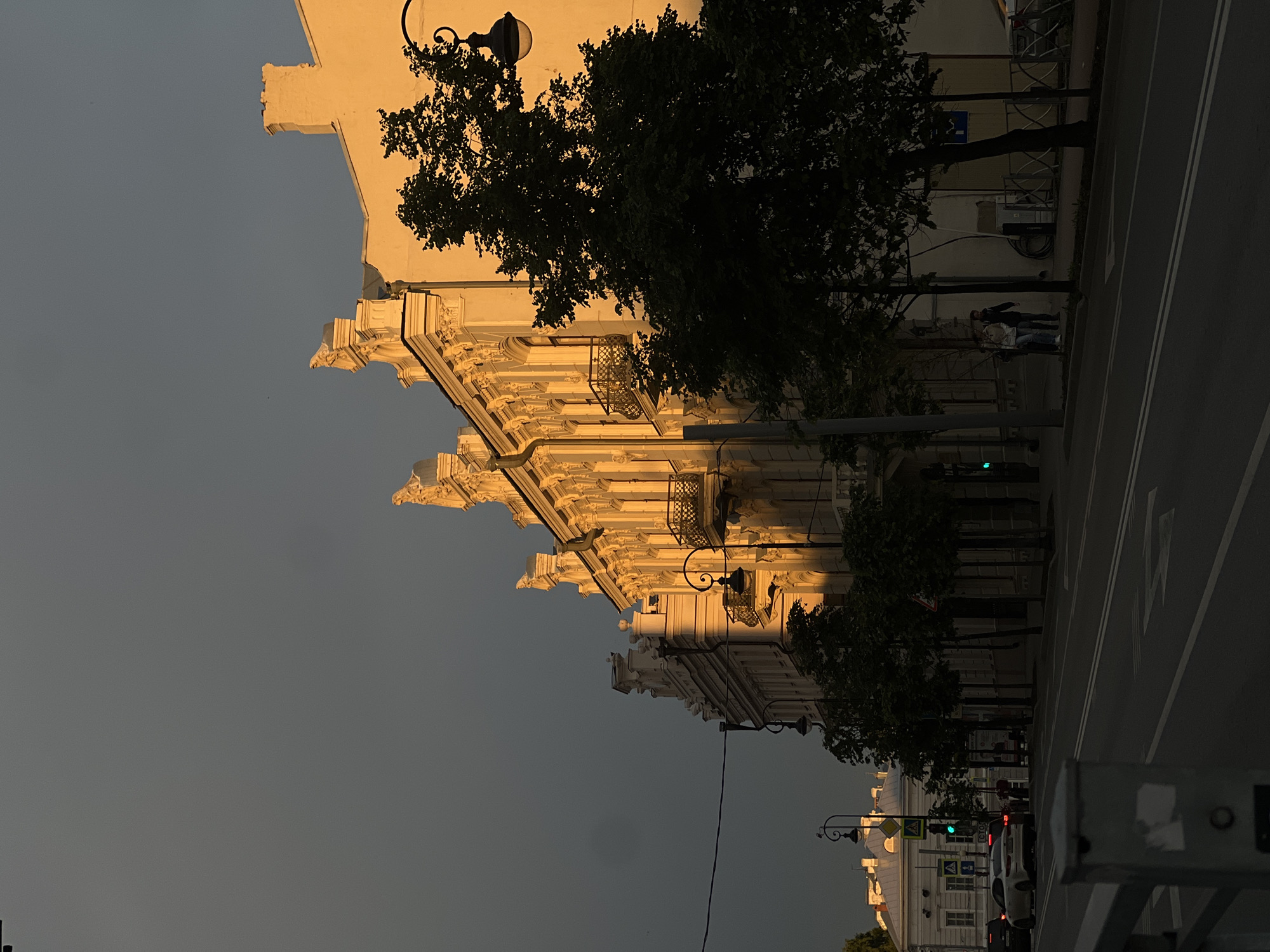 A vertical, eye-level shot shows a cityscape in Kazan, Russia, at dusk or dawn. A large, ornate building dominates the left side of the frame, illuminated by a warm, golden light. Its architectural details include balconies, decorative moldings, and protruding cornices. A tree partially obscures the building.
To the right is a street, marked by white dividing lines and containing automobiles. A few people are visible walking along the edge of the street, adjacent to the building. Street lamps line the side of the road. In the distance are more buildings under a similar golden light.
