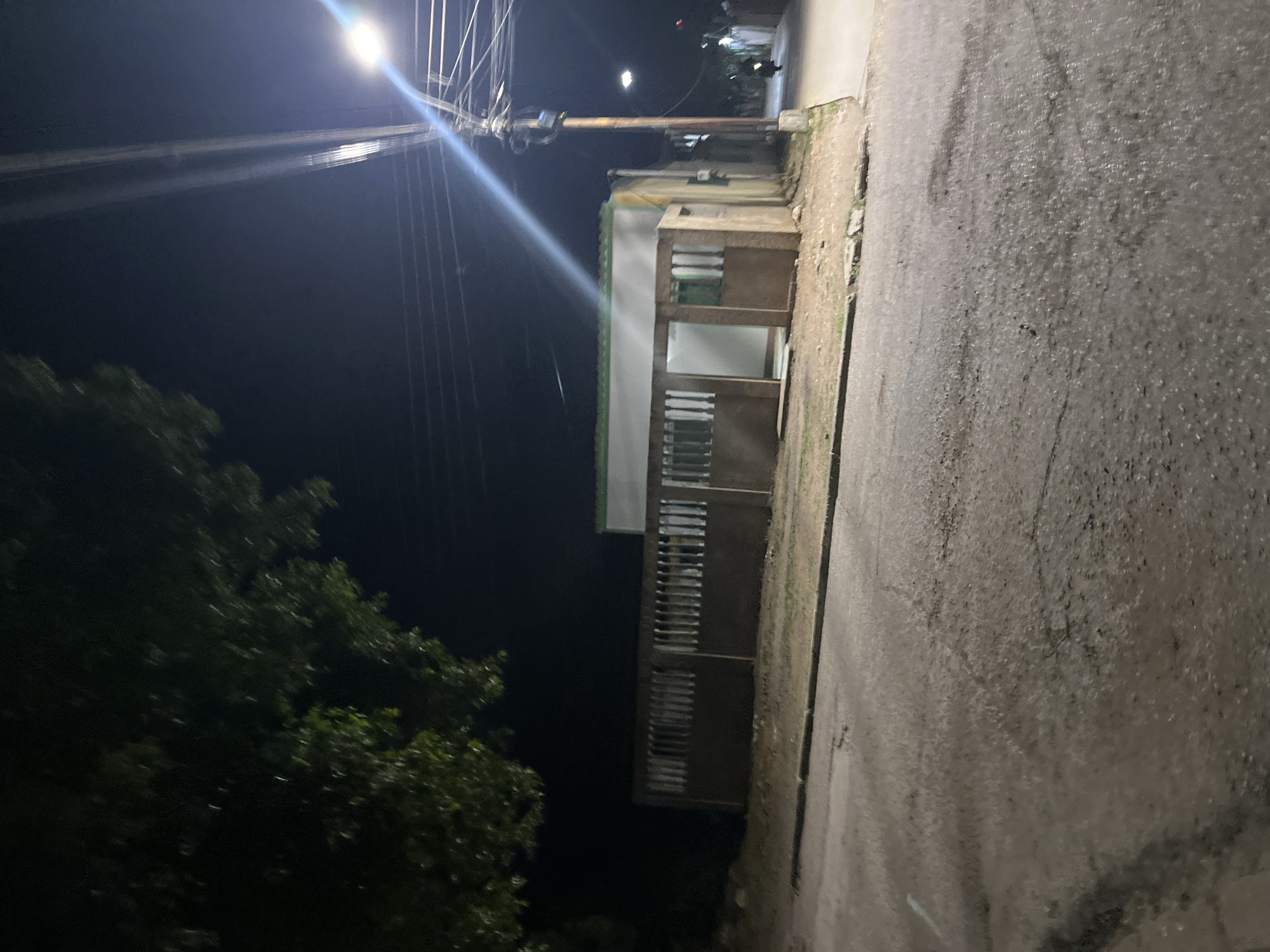 The image depicts a street scene at night in Santa Ana, Venezuela. A low-lying building with a light-colored facade and a green trim is visible, partially obscured by a concrete fence with vertical slats. The building appears to have an open entranceway. To the right, a utility pole with numerous wires and a bright street light illuminates the area. The street itself is paved and shows signs of wear, with some patches of lighter color suggesting repairs or puddles. A dark tree branches into the upper left corner of the frame. Further down the street, a distant light indicates other buildings, and a motorcycle with a rider is moving away from the camera. The overall atmosphere is dark and dimly lit, typical of a nighttime urban environment.