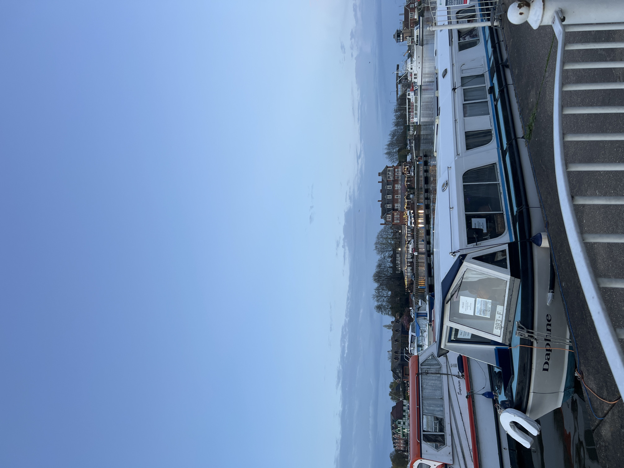 The image shows a waterfront scene in East Suffolk, United Kingdom. Several boats, including one named "Daphne", are docked along a concrete walkway. Behind the boats, buildings and trees line the shore. A white metal railing is visible on the right side of the image. The sky is a pale blue, indicating dusk or dawn. There are no people visible in the scene.