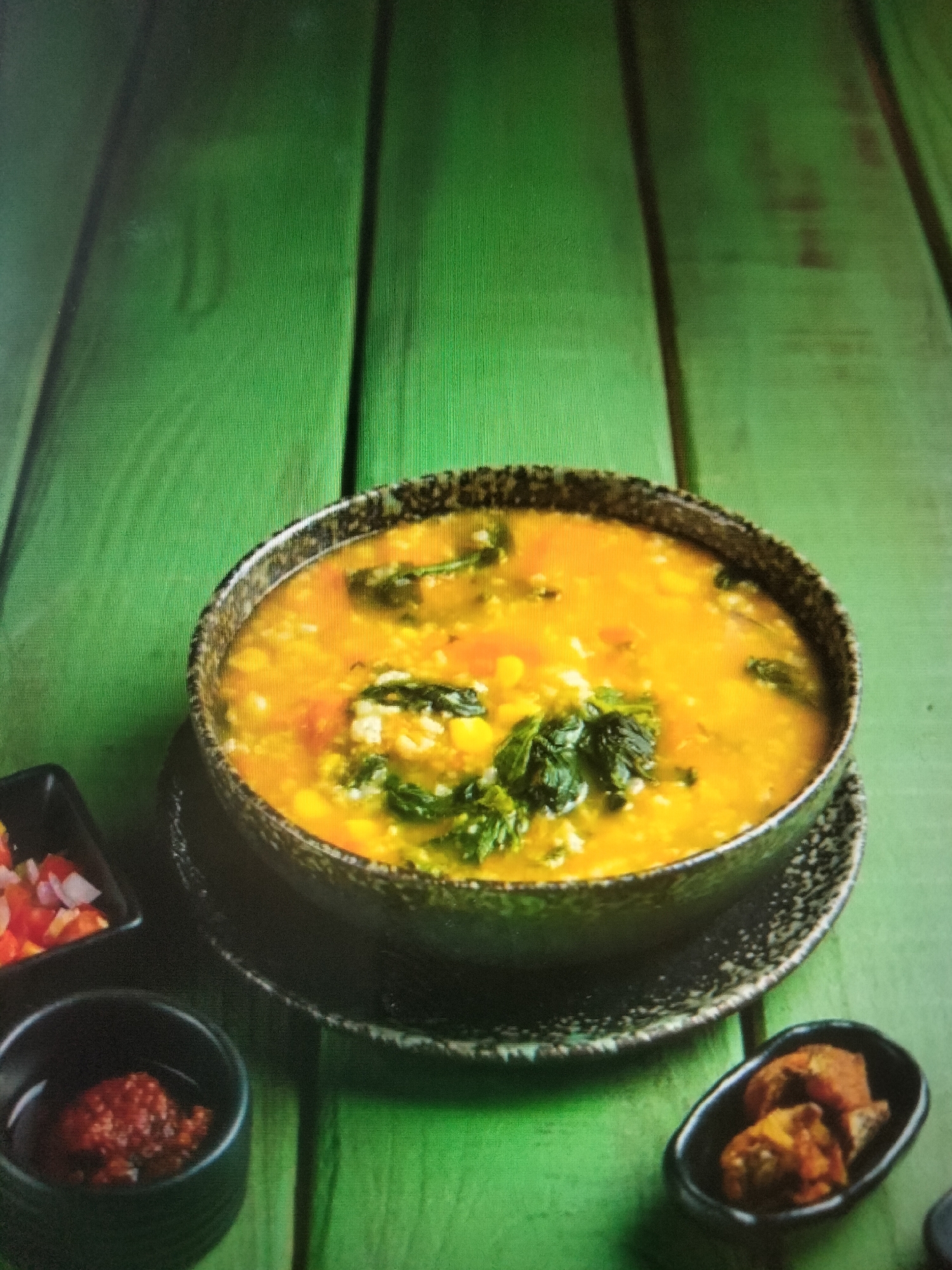 In the city of Payagaboh, Indonesia, a close-up, low-angle shot showcases a bowl of yellow soup with visible chunks of corn, spinach, and what appears to be rice or grains. The soup is placed on a dark, textured saucer which rests on a vibrant green wooden surface. To the left of the soup bowl, a small black dish contains finely diced red onions and tomatoes, appearing to be a side salad or salsa. Below and slightly to the left of that, another small black dish holds a dollop of a reddish paste, possibly a chili sauce or paste. To the right of the main soup bowl, a small black dish contains several pieces of fried food, possibly fish or meat. The green wooden surface is likely a table or a countertop, characterized by prominent vertical planks. The lighting appears to be overhead, casting soft shadows and highlighting the textures of the food and the wooden surface. There are no people visible in the image.
