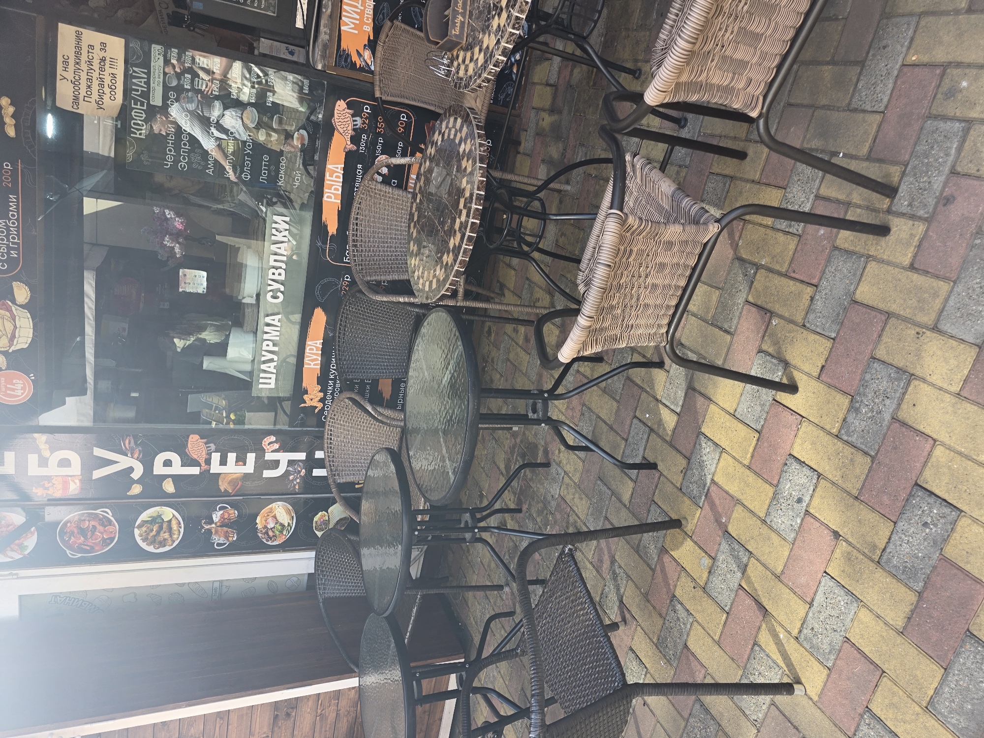 An outdoor cafe seating area is captured on a bright, sunny day, likely in Sochi, Russia. Several brown wicker chairs with metal frames are arranged around round, dark-framed tables, some with glass tops and one with a mosaic pattern. The ground is paved with interlocking bricks in patterns of red, yellow, and grey, casting shadows from the furniture.
Behind the seating area is a large glass storefront that reflects the sky and distant elements, suggesting an urban or semi-urban setting. Inside the reflection, a person can be vaguely seen. The storefront glass also displays numerous signs and menus, predominantly in Russian. A prominent sign reads: "У нас самообслуживание Пожалуйста убирайтесь за собой !!!" (We have self-service. Please clean up after yourselves !!!).
Other visible text includes a "КОФЕ/ЧАЙ" (Coffee/Tea) menu listing items like Черный кофе (Black coffee), Эспрессо (Espresso), Американо (Americano), Капучино (Cappuccino), Флэт Уайт (Flat White), Латте (Latte), Какао (Cocoa), and Чай (Tea) with various prices. A section for "ШАУРМА СУВЛАКИ" (Shawarma Souvlaki) is visible, along with menus for "КУРА" (Chicken) items (e.g., сердечки кури - chicken hearts) and "РЫБА" (Fish), specifying options like "стящая" and showing prices, for instance, "130гр 329р". A partially visible sign mentions "Tasty foc". The scene suggests a casual dining establishment open for business.