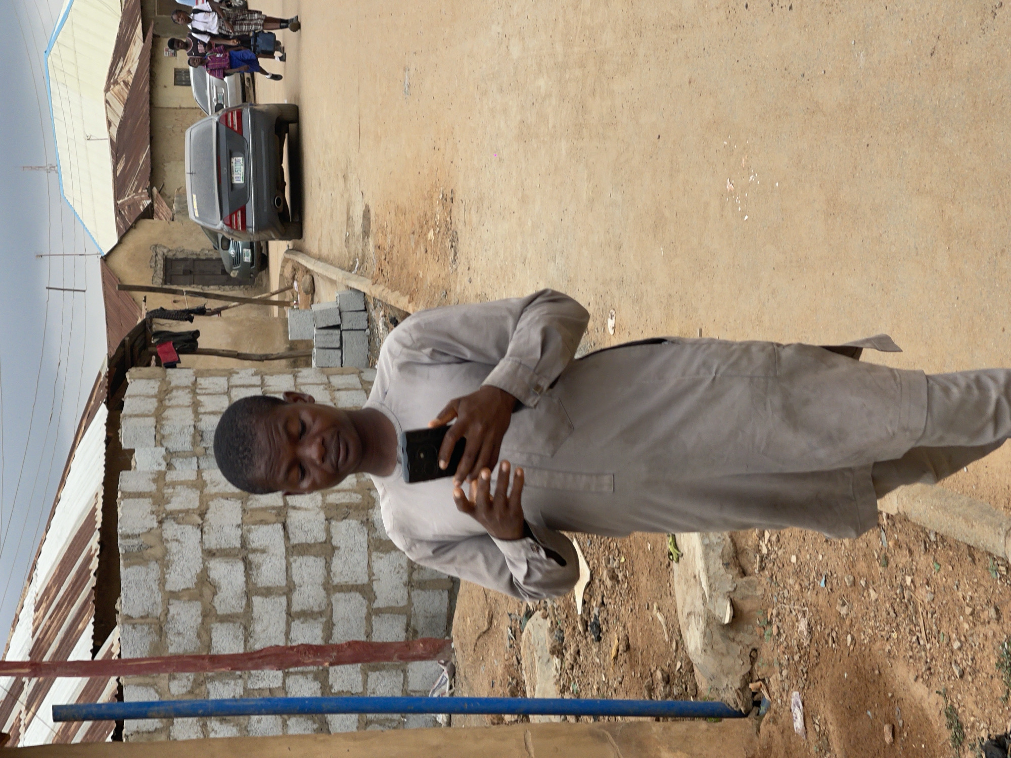 A man stands outdoors, looking at a cell phone he is holding in his hands. He is wearing a light-colored, long-sleeved garment. In the background, there is a partially constructed brick building with a corrugated metal roof. A sandy street runs alongside the building. In the distance, there are cars and pedestrians. The location appears to be a residential area. The image may be in the Karmajiji area of Nigeria.
