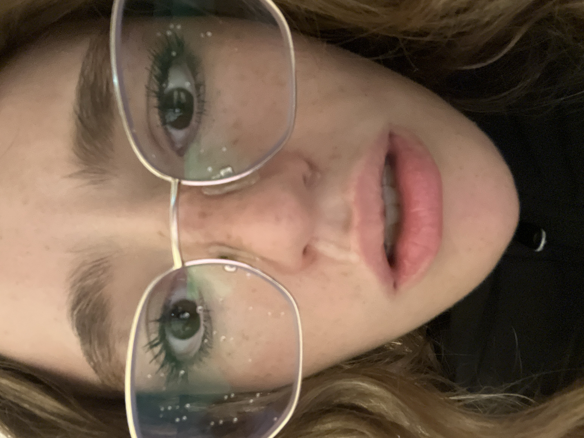 A close-up, rotated image shows a person's face, partially obscured by eyeglasses with water droplets on the lenses. The person has fair skin with freckles. Their eyes are green and eyebrows are light brown. The person's lips are a pinkish-red. Hair is light brown and slightly wavy. A dark fabric or background is visible on the right side of the image. The location, San Bernardino Tlaxcalancingo, Mexico, is not discernible from the image.
