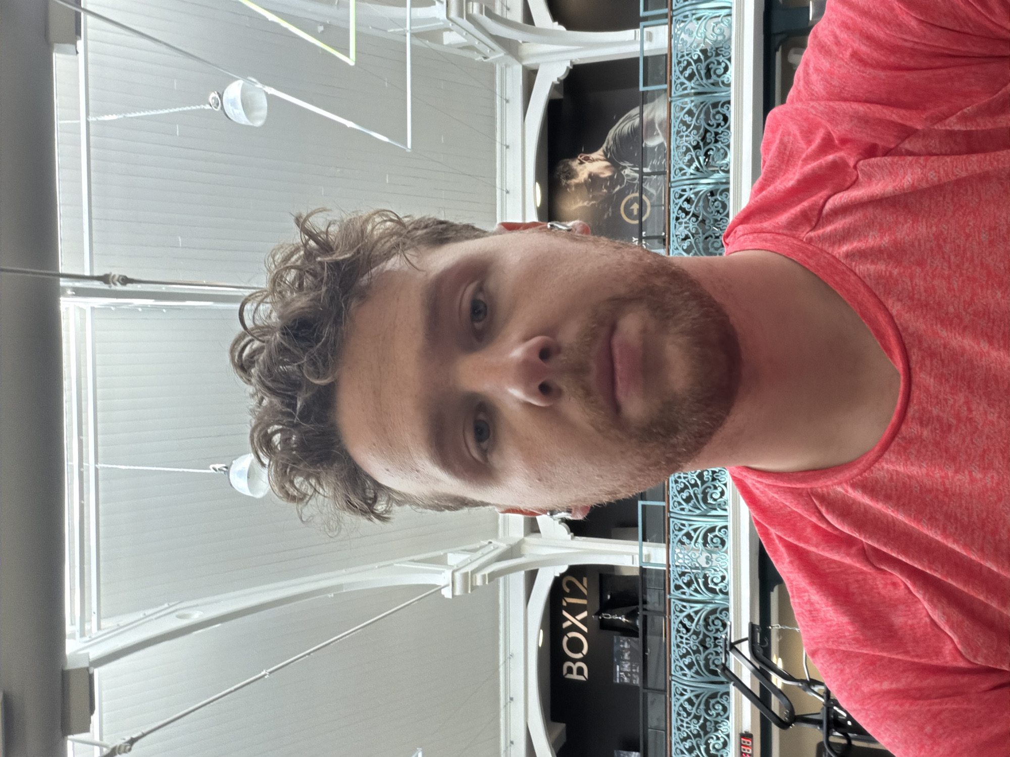 A close-up, slightly angled shot features a person with curly hair and a beard, wearing a red t-shirt. The background reveals elements of an interior space. Architectural details are visible, with white structural supports and modern lighting fixtures hanging from the ceiling. There is also an advertisement for BOX12. The setting appears to be a public space in London, United Kingdom, as indicated by the style of architecture and the shop's name.