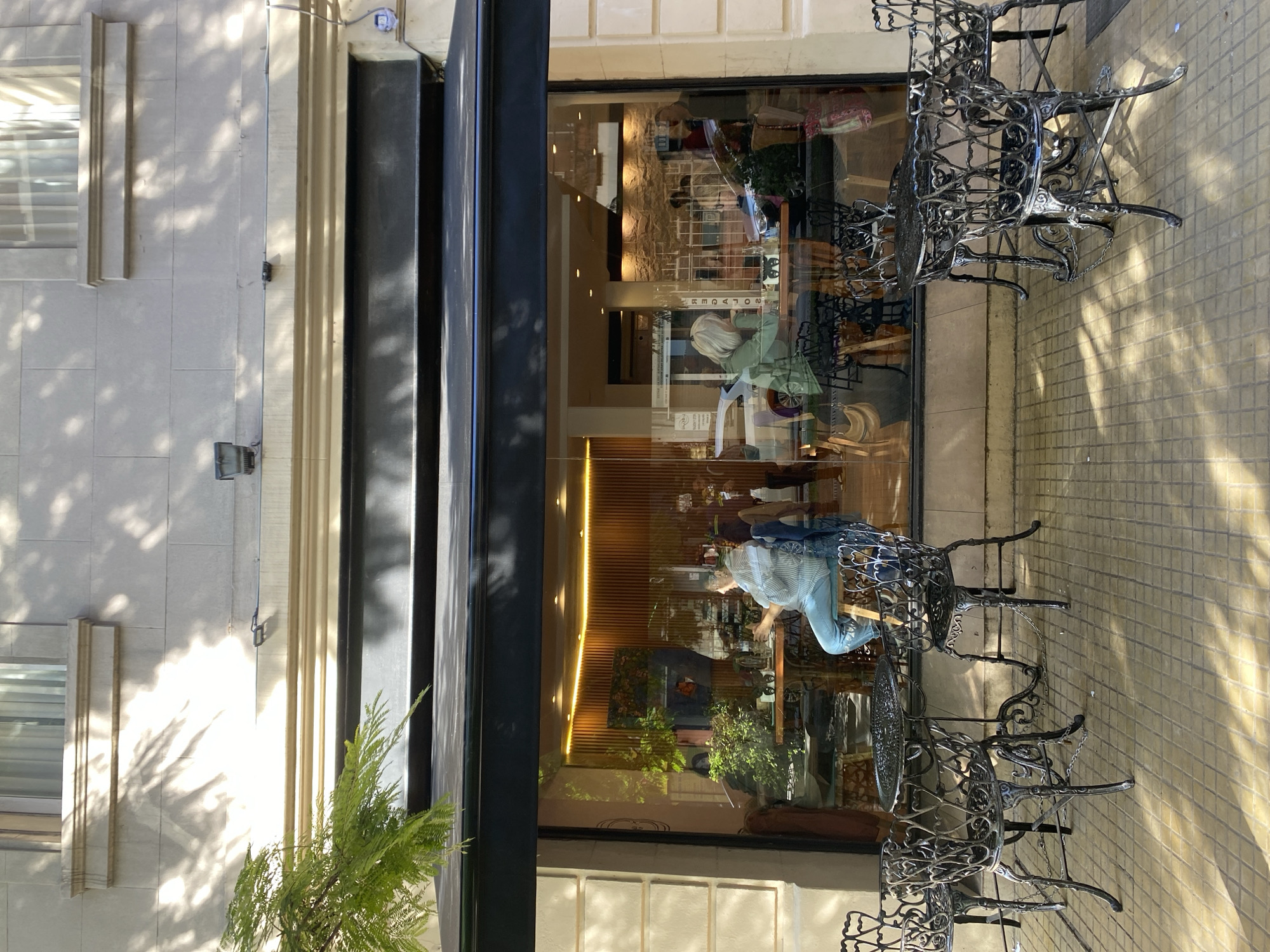 The image captures the exterior of a shop or cafe in Buenos Aires, Argentina. A black awning shades the entrance. Through the large glass window, the interior is visible, showing tables and chairs, as well as a woman with blonde hair sitting and looking down at the table. The interior features warm lighting from vertical light strips. Outside the shop, black wrought iron tables and chairs are arranged on the sidewalk. The sidewalk is paved with light-colored square tiles. A building facade with light-colored stone is to the left of the shop. The overall scene suggests a bright, sunny day with dappled sunlight casting shadows on the building and sidewalk.