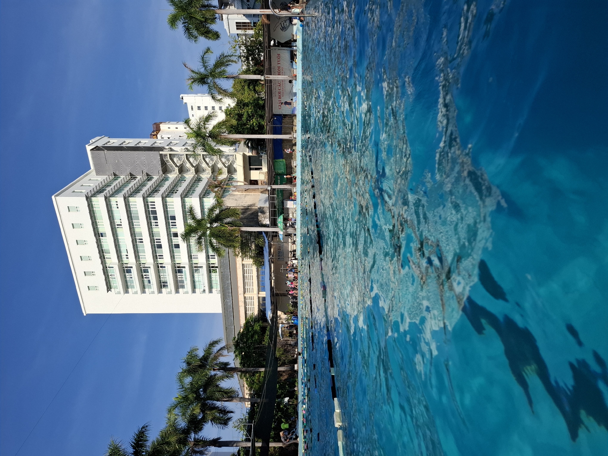 The image captures a sunny day at a swimming pool in Nha Trang Ward, Vietnam. The foreground is dominated by the clear, bright blue water of the pool, with visible ripples and reflections of the sky and surrounding structures. Floating lane markers, black and white with yellow buoys, divide the pool into swimming lanes. In the middle ground, palm trees line the edge of the pool, with a group of children and adults standing near the pool's edge, some appearing to be participating in or observing an activity. Behind them, a building with a white facade, multiple floors, and numerous windows stands prominently against a clear blue sky. The architecture suggests a modern, multi-story structure. In the background, other buildings and trees are visible, contributing to an urban, yet somewhat tropical, environment. A banner with Vietnamese text is visible on a fence behind the pool area, indicating a local event or establishment. The overall scene suggests a public recreational facility, likely a school or community pool, given the presence of children.