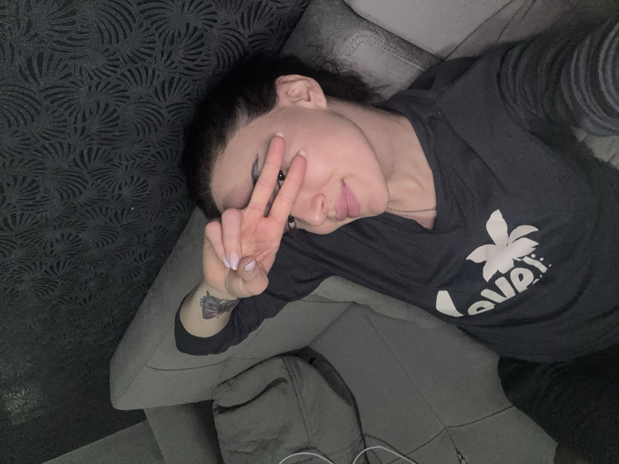 A person is lying on a grey upholstered sofa, making a peace sign with their right hand. The person has dark hair and is wearing a dark long-sleeved shirt with a white design. A tattoo is visible on their right wrist. The sofa is positioned against a textured dark wallpaper with a repeating pattern. The location cannot be determined from the image.