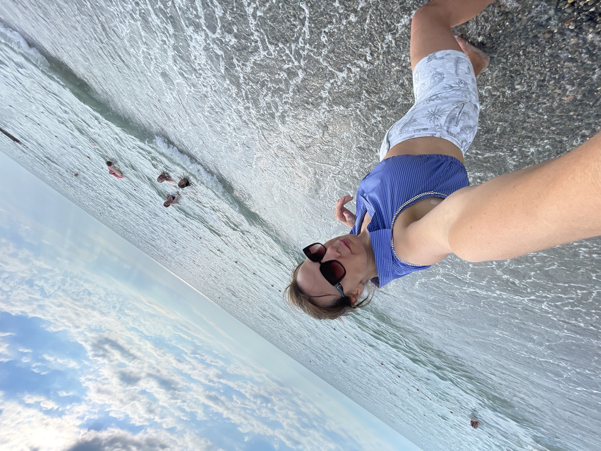 In a selfie taken at sea level, a person stands near the water's edge, facing the camera. They are wearing a blue and white striped button-down shirt and patterned shorts, with sunglasses perched on their nose. The person appears to be looking at the camera with a slight smile. The background is a mix of the ocean and the sky, with soft clouds visible. In the distance, other people can be seen in the water. The scene suggests a casual day at the beach, possibly in Sochi, Russia, given the context. The lighting indicates it is daytime, and the weather appears pleasant, with clear skies and calm water. The overall mood is relaxed and summery. No visible text is present in the image.