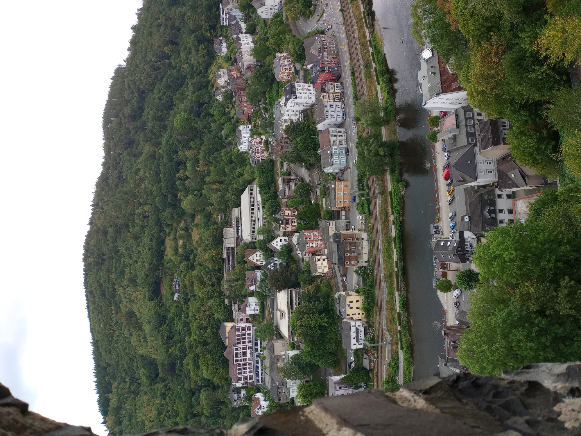The image shows a panoramic view of the city of Altena, Germany, from a high vantage point, possibly a castle or tower. The foreground is dominated by the rough, textured stone of a wall, framing the scene.
In the mid-ground, a dense, verdant forest covers a steep hillside, with a cluster of buildings nestled at its base. These buildings are a mix of architectural styles, with some featuring red-tiled roofs and traditional facades, while others are more modern, with white and grey exteriors. A river flows along the bottom of the hillside, bordered by a road with several parked cars. The river appears calm, with subtle reflections on its surface.
Further into the background, more buildings are visible, some larger and more institutional-looking, possibly schools or administrative buildings, interspersed with residential houses. The dense green foliage of the forest continues to dominate the upper portion of the image, extending up to the horizon line. The sky is overcast, contributing to a muted, natural light. There are no visible people in the frame. The overall impression is one of a picturesque European town nestled within a natural landscape.