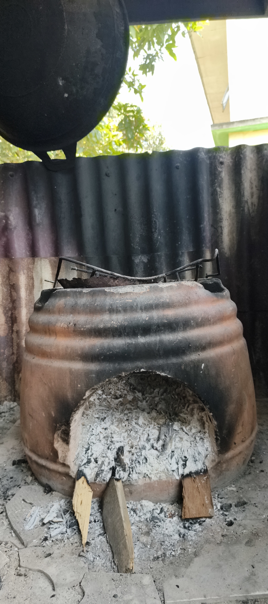 In Muaro Jambi, Indonesia, an outdoor cooking scene features a traditional clay stove, possibly used for preparing food over an open fire. The stove is a large, round, terracotta-colored vessel with a wide opening at the front, filled with white ash and two pieces of light-colored wood, suggesting recent use. A rusty metal grate sits on top of the stove, likely to hold a cooking pot. Behind the stove, a corrugated metal barrier, blackened by soot, partially encloses the area. In the background, lush green foliage and sunlight filter through, with a hint of a building structure visible. The ground is paved with light-colored tiles, scattered with more ash.
