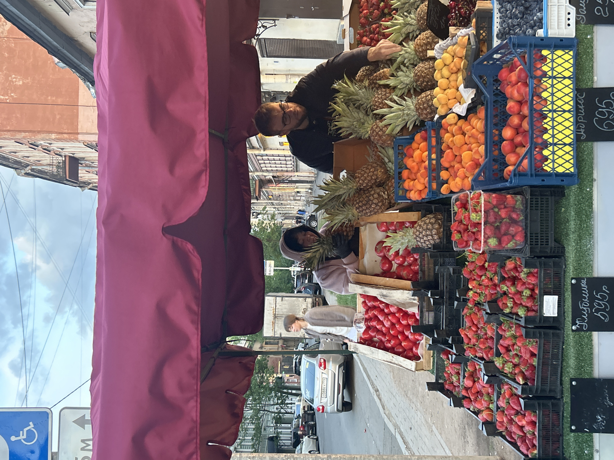 This image captures an outdoor fruit stall on a city street in Saint Petersburg, Russia. The scene features a variety of fresh produce displayed under a reddish-purple canopy.
A man with glasses and a beard, wearing a dark top, is actively tending the stall, reaching into a large pile of pineapples. Another individual, wearing a light-colored head covering, is also visible under the canopy, seemingly arranging or selecting fruit. A third person walks on the sidewalk in the background, partially obscured.
The stall showcases numerous types of fruit, including abundant pineapples, multiple crates of bright red strawberries, and baskets filled with apricots or peaches. Blueberries or cherries are also visible in a blue crate. The fruits are neatly presented in a mix of black plastic, blue plastic, and wooden crates, arranged on display stands along the pavement.
Visible text on chalkboards indicates prices and fruit types in Cyrillic. One sign clearly reads "Клубника" (Strawberry) with "595р" (595 rubles), and another displays "Абрикос" (Apricot) with "695" (695 rubles). A partial price of "25" is also discernible. A faint sign suggests market operating hours "8.00-20.00".
The setting is an urban environment characterized by multi-story buildings with classic European architecture, lining the street. Several cars are parked along the curb, including a silver vehicle with a Russian license plate "C088BY 174". Overhead power lines crisscross against a partly cloudy sky. A blue sign indicating disabled access and a white arrow sign are visible in the foreground. The scene depicts a lively daytime market, likely in the late morning or afternoon, under clear, natural light.