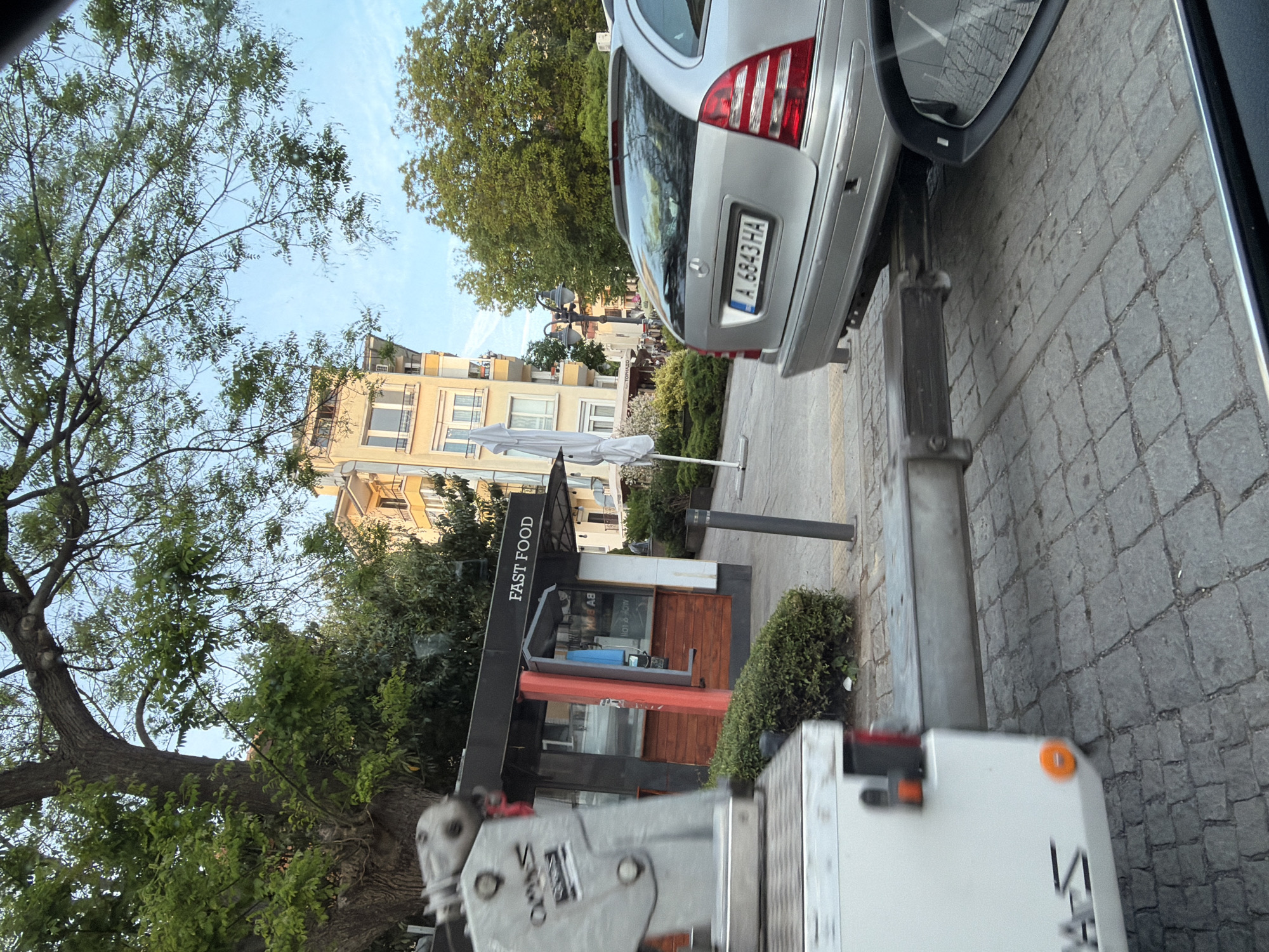 The image shows a street scene in Burgas, Bulgaria. A silver Mercedes-Benz car with license plate "A 6843 HA" is parked on a cobblestone street. Next to the car is a "FAST FOOD" kiosk with a wooden facade. A tree is positioned on the left side of the image, with apartment buildings visible in the background. A service vehicle is located in the foreground, partially obstructing the view.