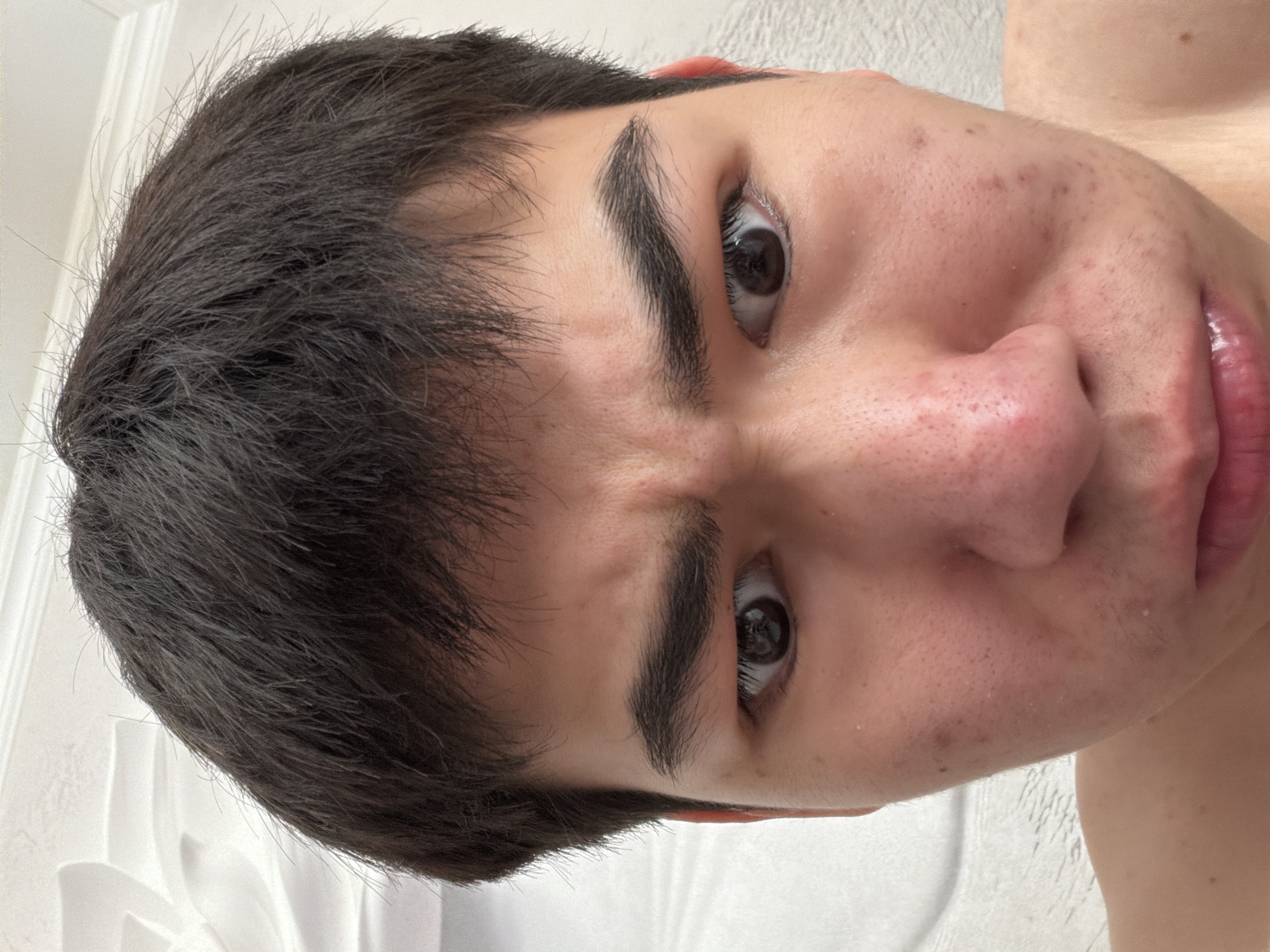 The image is a close-up, selfie-style photograph of a young person's face. The subject has dark, messy hair and is looking directly at the camera with a furrowed brow and a slightly downturned mouth, suggesting a displeased or concerned expression. Their skin appears to have some texture and minor blemishes. The background is out of focus but appears to be a textured white surface, possibly a wall or ceiling with some decorative elements. There is no indication of the city of Shymkent, Kazakhstan, or any urban environment within the image.
