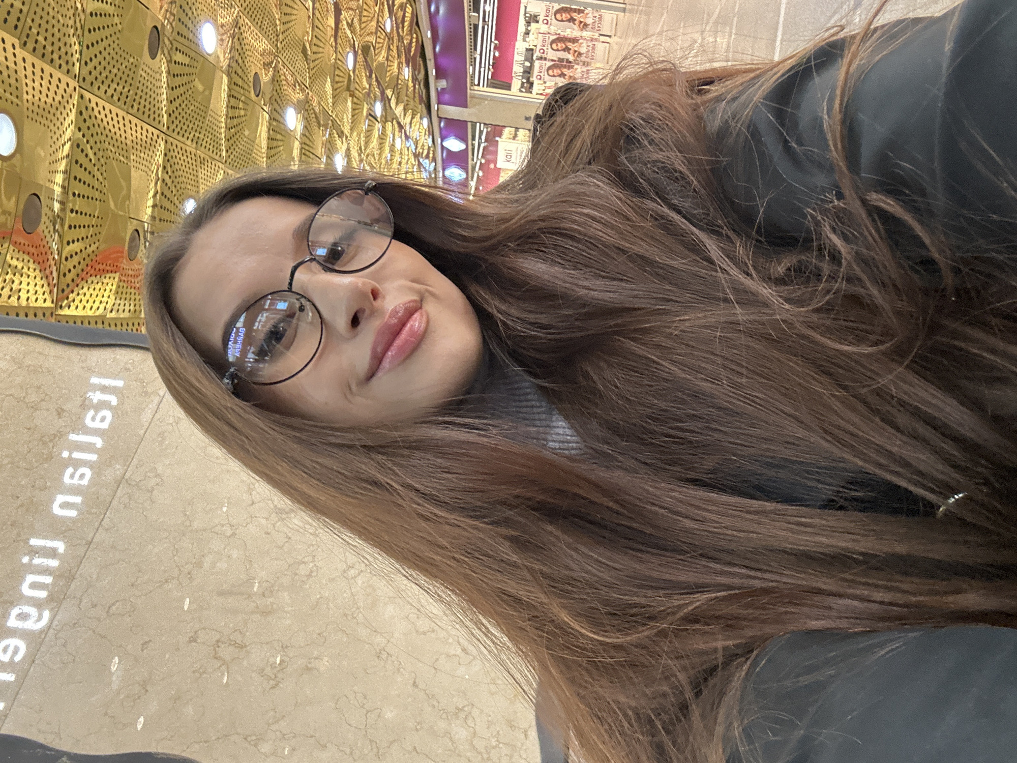 A woman with long, brown hair, wearing glasses, is posing for a selfie. She appears to be indoors, in a brightly lit area. The background shows a distinctive patterned wall and other retail displays, suggesting a shopping mall or similar commercial space. The floor has a light-colored, speckled pattern. A partially visible sign in Russian, "линии линдт," indicates the location is likely in Russia. Given the retail context and the Russian signage, a likely location would be in a mall within the city of Yekaterinburg, Russia.