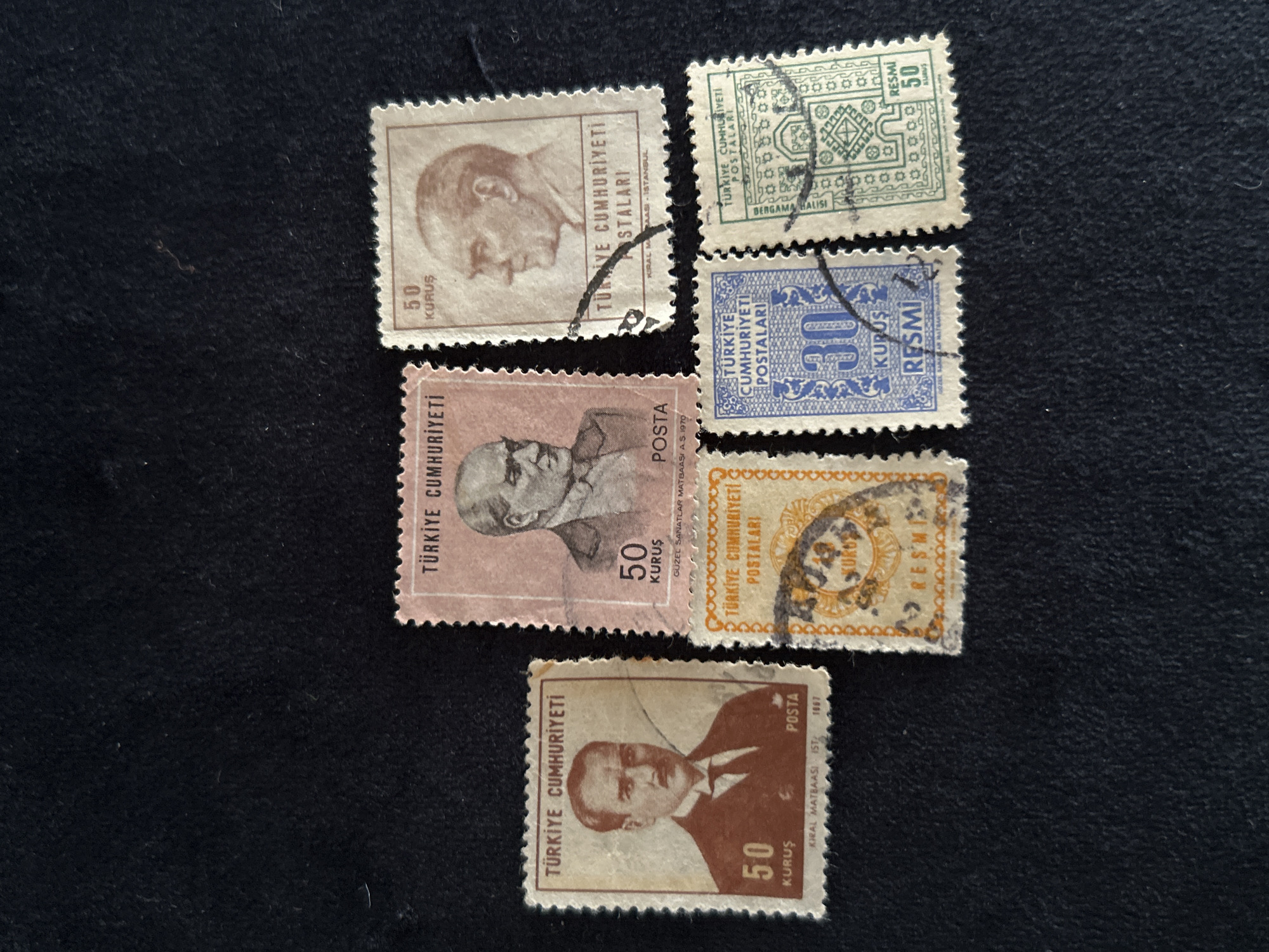 Six Turkish postage stamps are displayed on a dark, textured surface. The stamps are arranged in a roughly horizontal line, with some overlap.
From left to right:
The first stamp is light brown with a portrait of a man in a suit and tie. "TÜRKİYE CUMHURİYETİ" is printed at the top, and "50 KURUŞ POSTA" is at the bottom. Below that, "KİRAL MATBAASI IST. 1967" is visible.
The second stamp is pinkish with a portrait of a man in a suit and tie, facing right. "TÜRKİYE CUMHURİYETİ" is at the top. "50 KURUŞ" and "POSTA" are below the portrait. "GÜZEL SANATLAR MATBAASI A.Ş. 1970" is at the very bottom.
The third stamp is also pinkish with a portrait of a man in a suit and tie, facing left. "TÜRKİYE CUMHURİYETİ" is at the top. "50 KURUŞ" and "POSTALARI" are below the portrait. "KİRAL MATBAASI - İSTANBUL" is at the bottom.
The fourth stamp is light orange and has a circular design with text. "TÜRKİYE CUMHURİYETİ POSTALARI" is visible. "10 KURUŞ RESMİ" is printed below the circular design.
The fifth stamp is blue with a large "30" in the center, surrounded by decorative elements. "TÜRKİYE CUMHURİYETİ POSTALARI" is at the top. "KURUŞ RESMİ" is at the bottom.
The sixth stamp is light green with a geometric pattern. "TÜRKİYE CUMHURİYETİ POSTALARI" is at the top. "BERĞAMA HALISI" is printed vertically on the left side. "RESMİ 50 KURUŞ" is at the bottom right.
