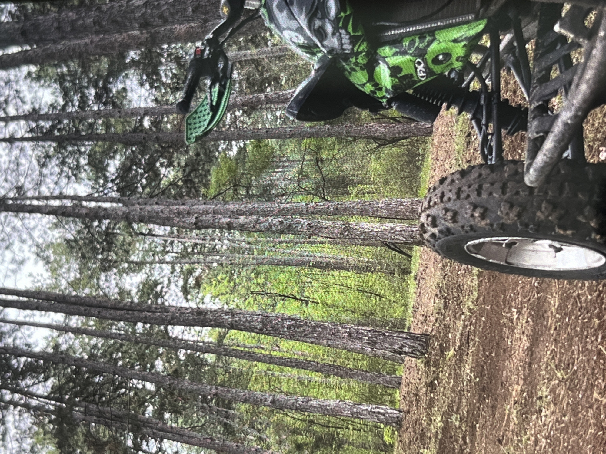 The image shows a close-up of an off-road vehicle, possibly an ATV, in a wooded area. The vehicle is positioned on a dirt or grassy terrain. The front wheel and part of the handlebars are visible, along with some green and black camouflage-patterned bodywork. The tire is muddy, suggesting recent use. The background consists of a dense forest of tall, thin trees. The location of the scene is Unknown, Unknown.