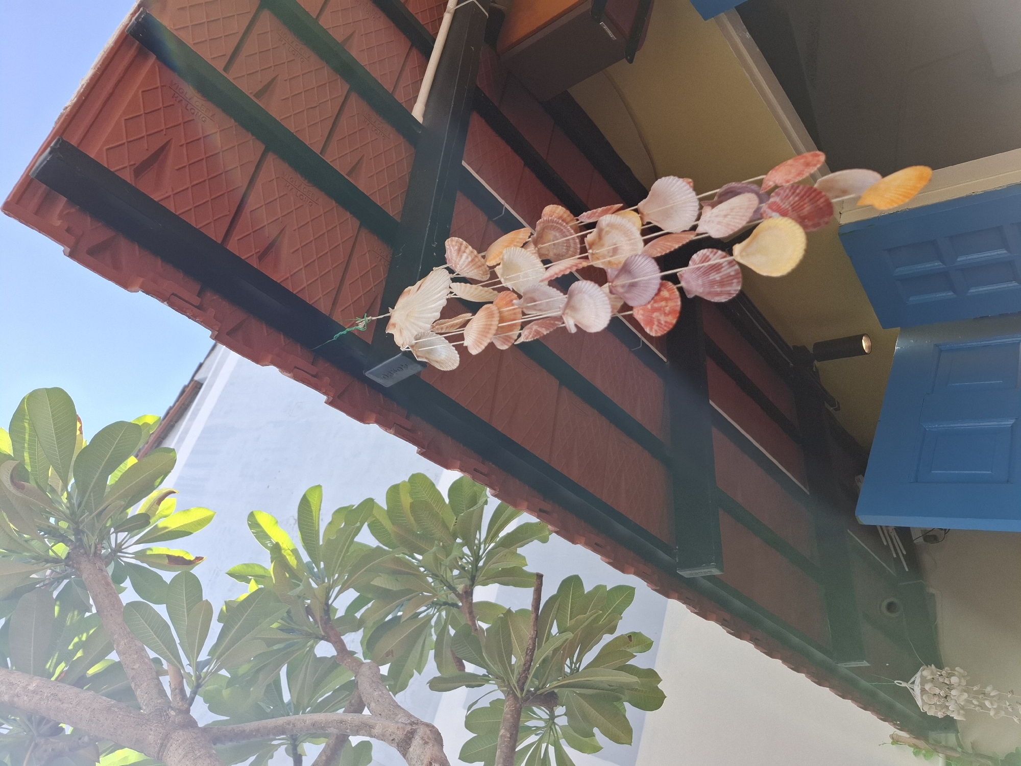 An outdoor view of the roofline of a building with red tile roofing and exposed dark green beams. A string of seashell wind chimes hangs from the roof. To the left, a tree with large green leaves is visible, with sunlight filtering through the foliage. The sky is clear and blue. Below the roofline, the top of a bright blue door is visible. The scene appears to be in Nha Trang, Vietnam, suggested by the architectural style and the presence of tropical foliage. No people are present.